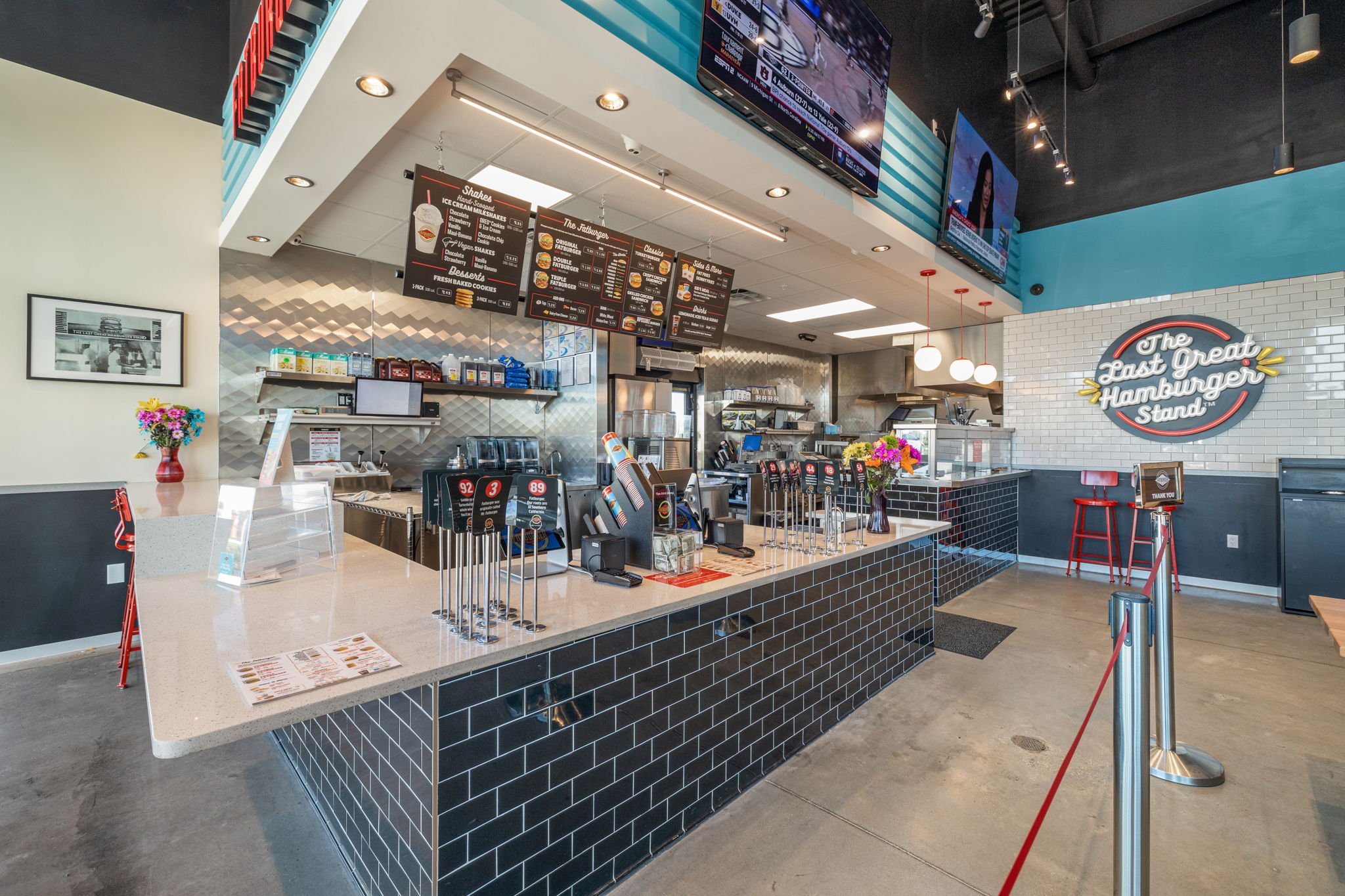 Interior of a Fatburger fast-food burger restaurant with a counter, menu boards, multiple digital screens, and a sign saying 'The Last Great Hamburger Stand.' Bright lighting and clean design.
