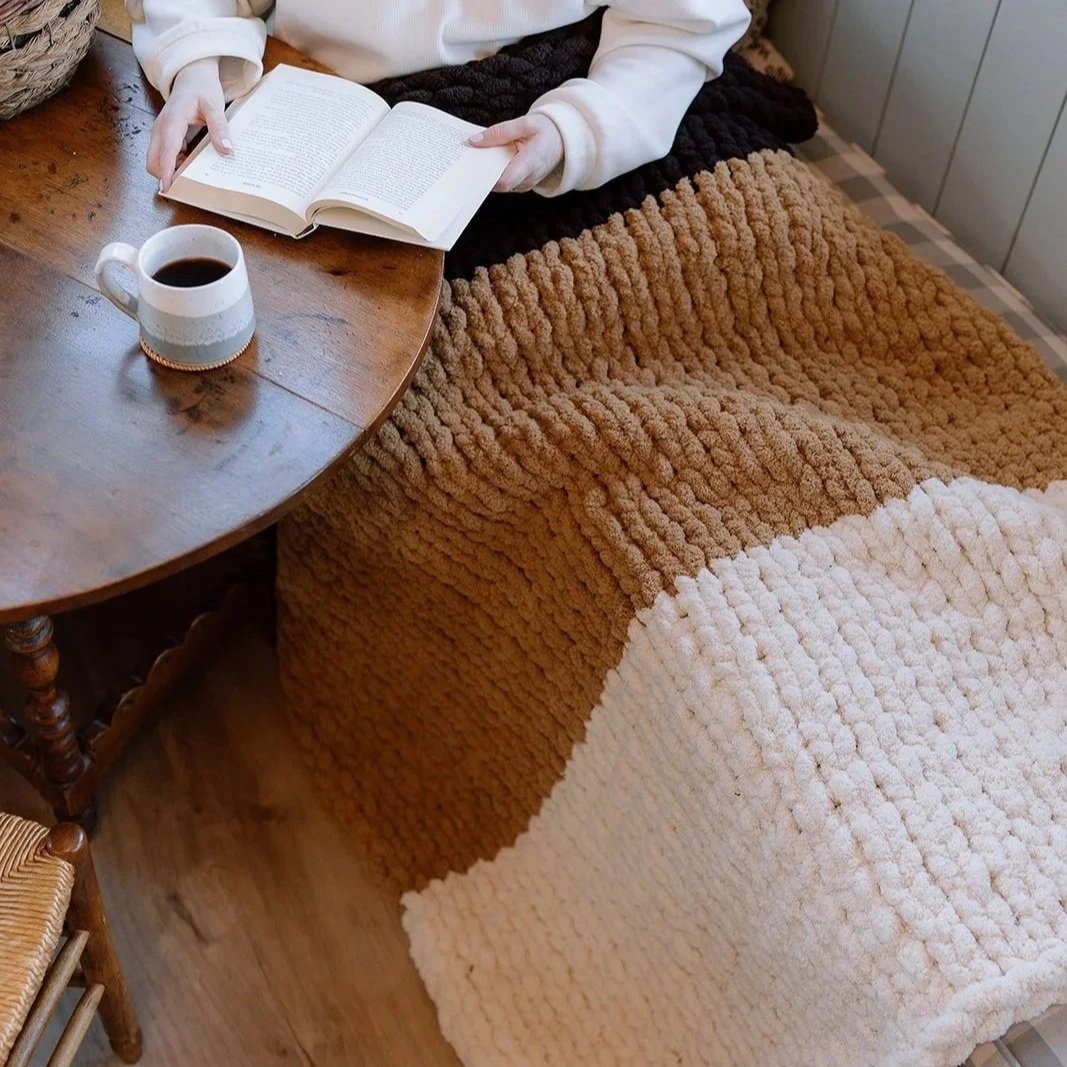 Person reading a book at a wooden table with a cup of coffee, covered by a chunky knit blanket in brown, black, and cream colors.