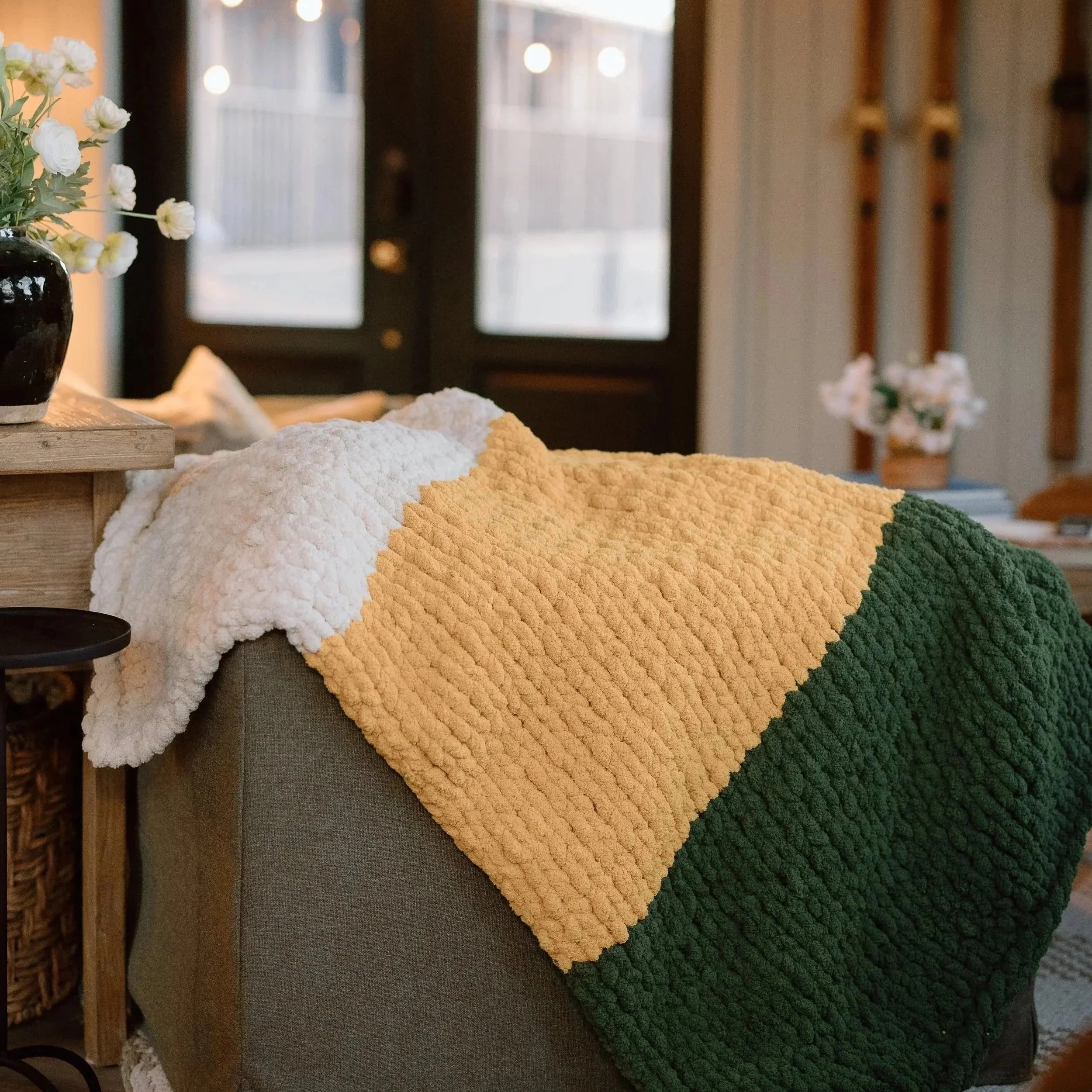 Colorful knitted blankets in white, yellow, and green draped over the arm of a gray sofa in a cozy living room, with a wooden table with a black vase of white flowers nearby and a wooden staircase in the background. luxury hand knit blanket