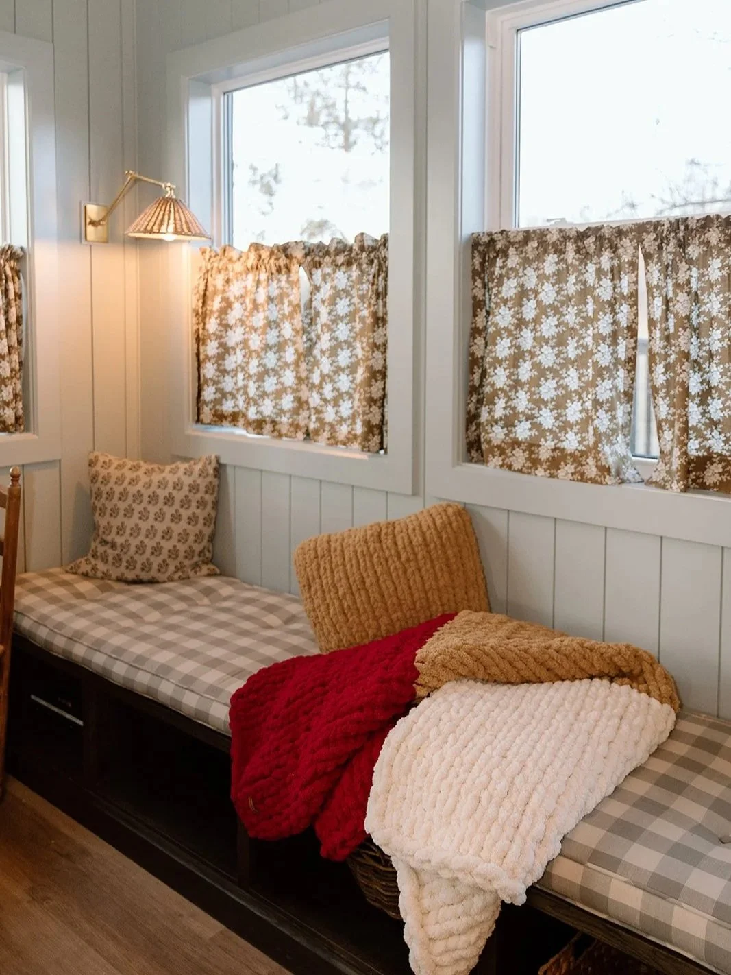 A cozy corner with a window seat featuring checkered and floral pillows, and blankets in red, beige, and white. The window has floral curtains, and there's a wall-mounted lamp with a bronze finish.