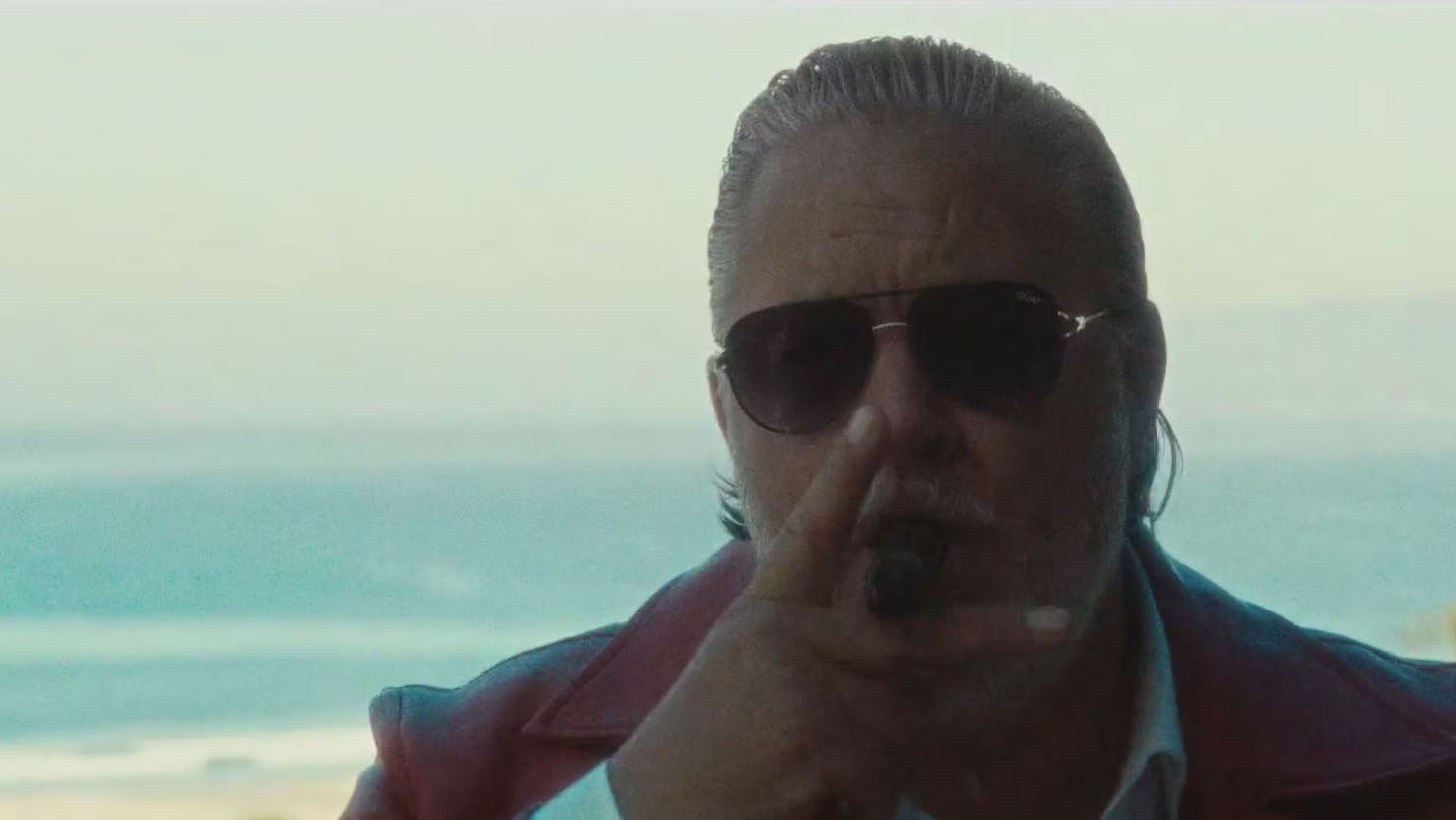 Man wearing sunglasses and red jacket, smoking a cigar with a beach view in the background.