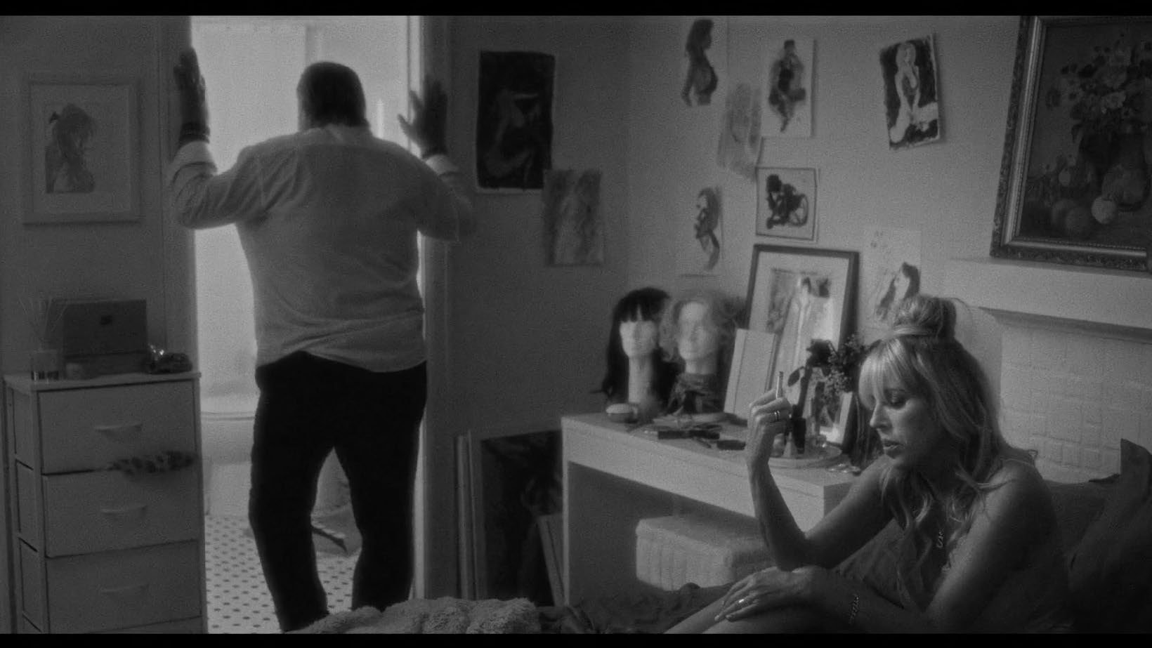 Black and white photo showing a woman sitting on a bed, holding a cigarette, looking pensive. The room is decorated with framed artwork and wigs on mannequins. A man stands by the doorway.