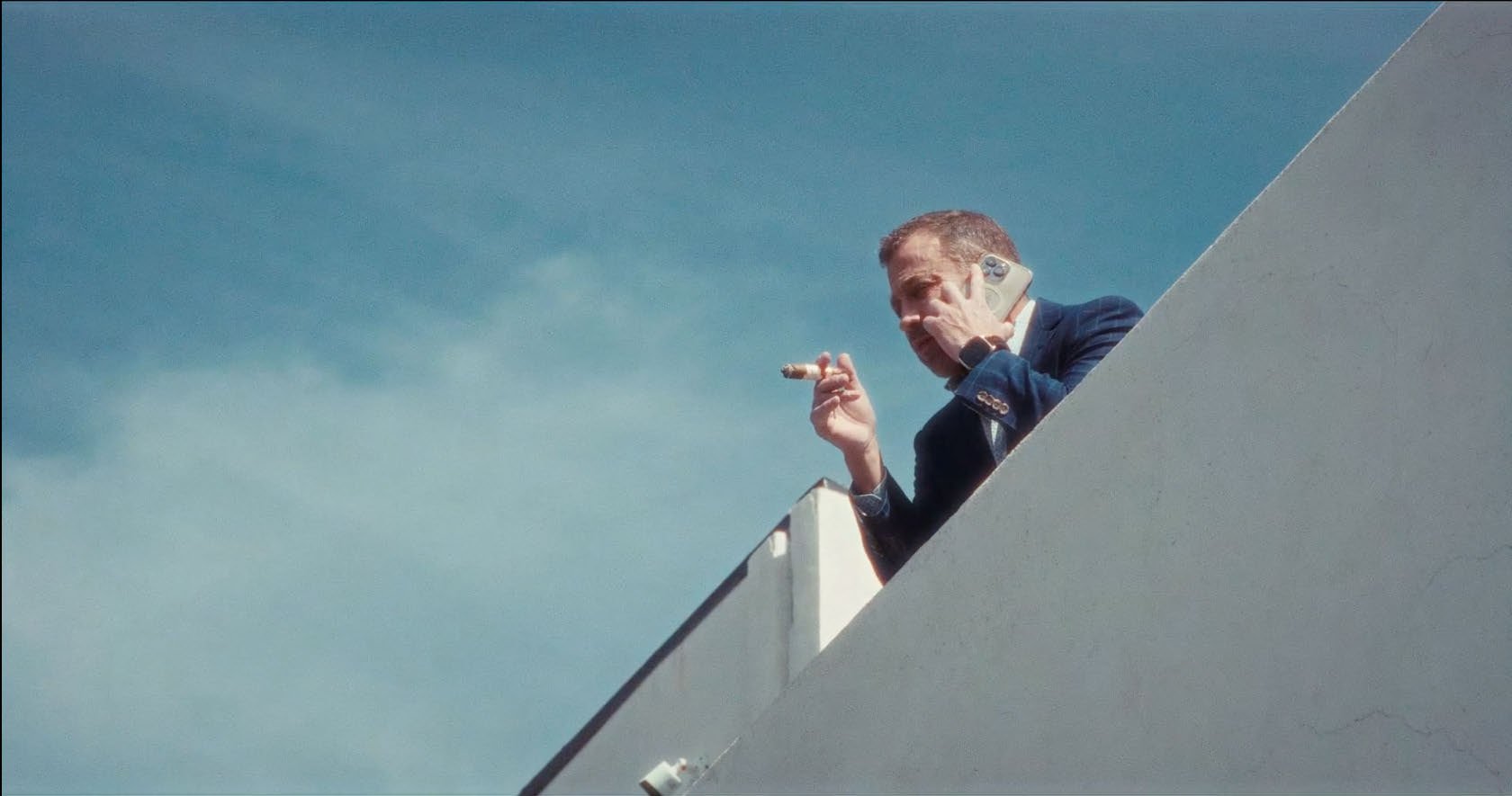 Man in suit on balcony holding phone and cigar against clear sky.