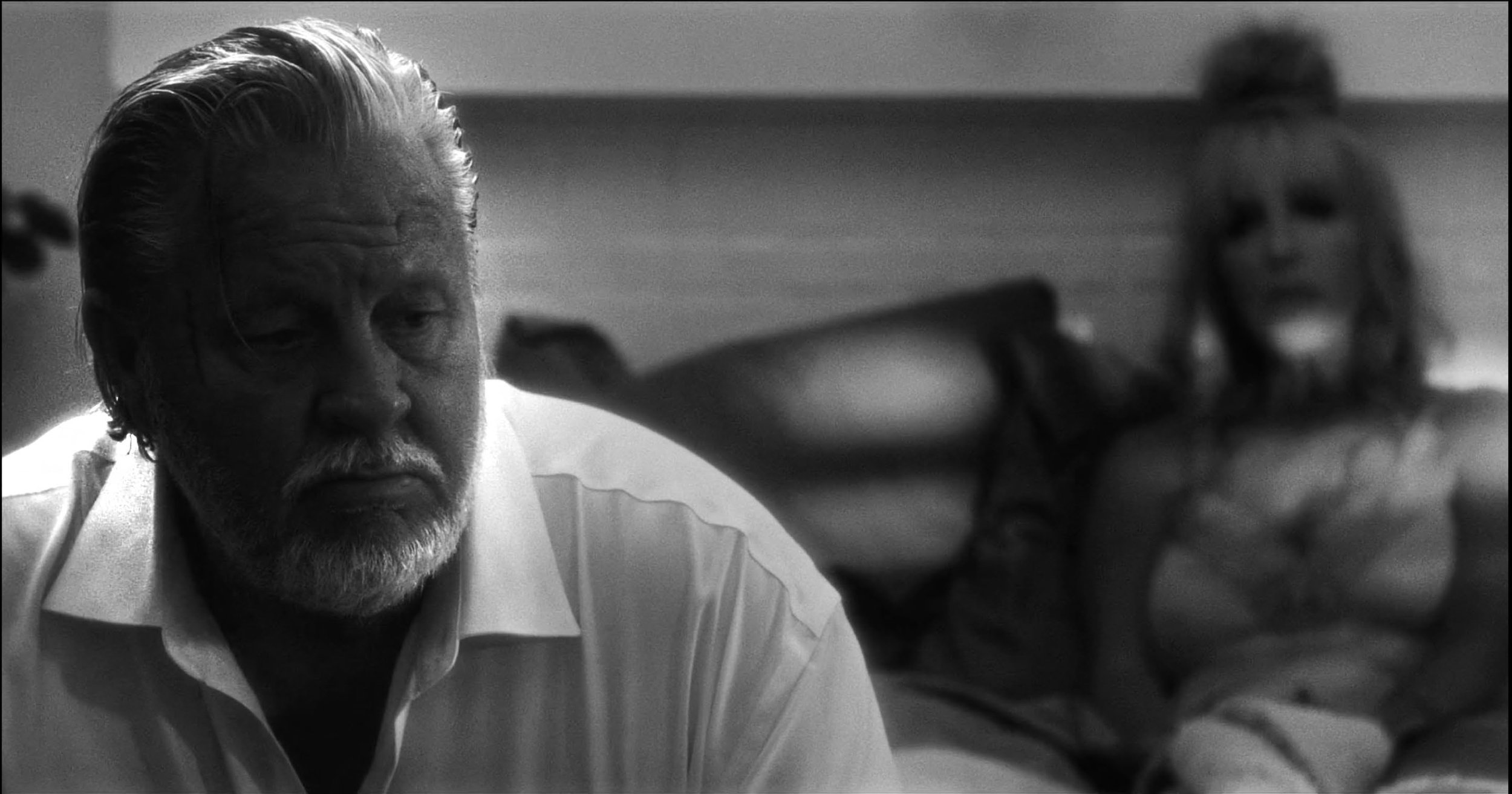 Black and white image of an elderly man in focus sitting in the foreground, with a blurred woman in the background sitting on a bed.