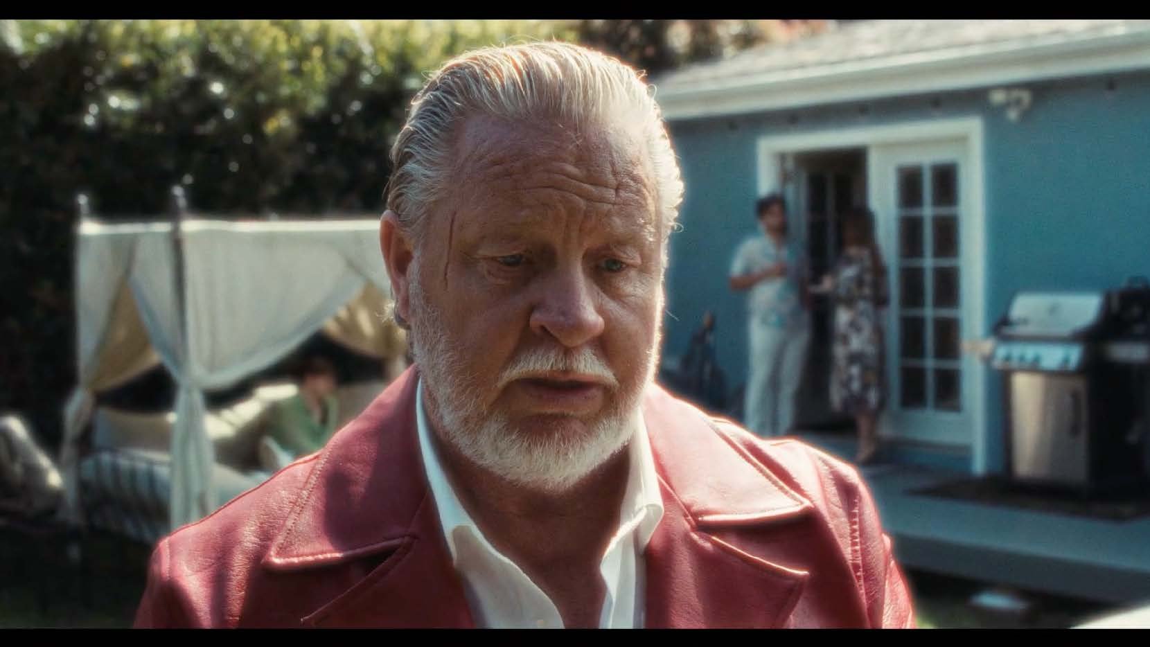 A man with a white beard and slicked-back hair in a red jacket is in the foreground. In the background, there is a blue house with a barbecue grill, and two people standing by a door.