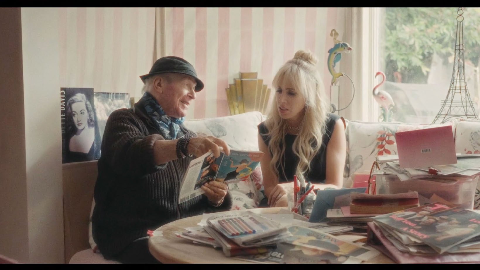 Two people sitting at a cluttered table with magazines and books, discussing a book. The room has decorative items like a flamingo and an Eiffel Tower replica.