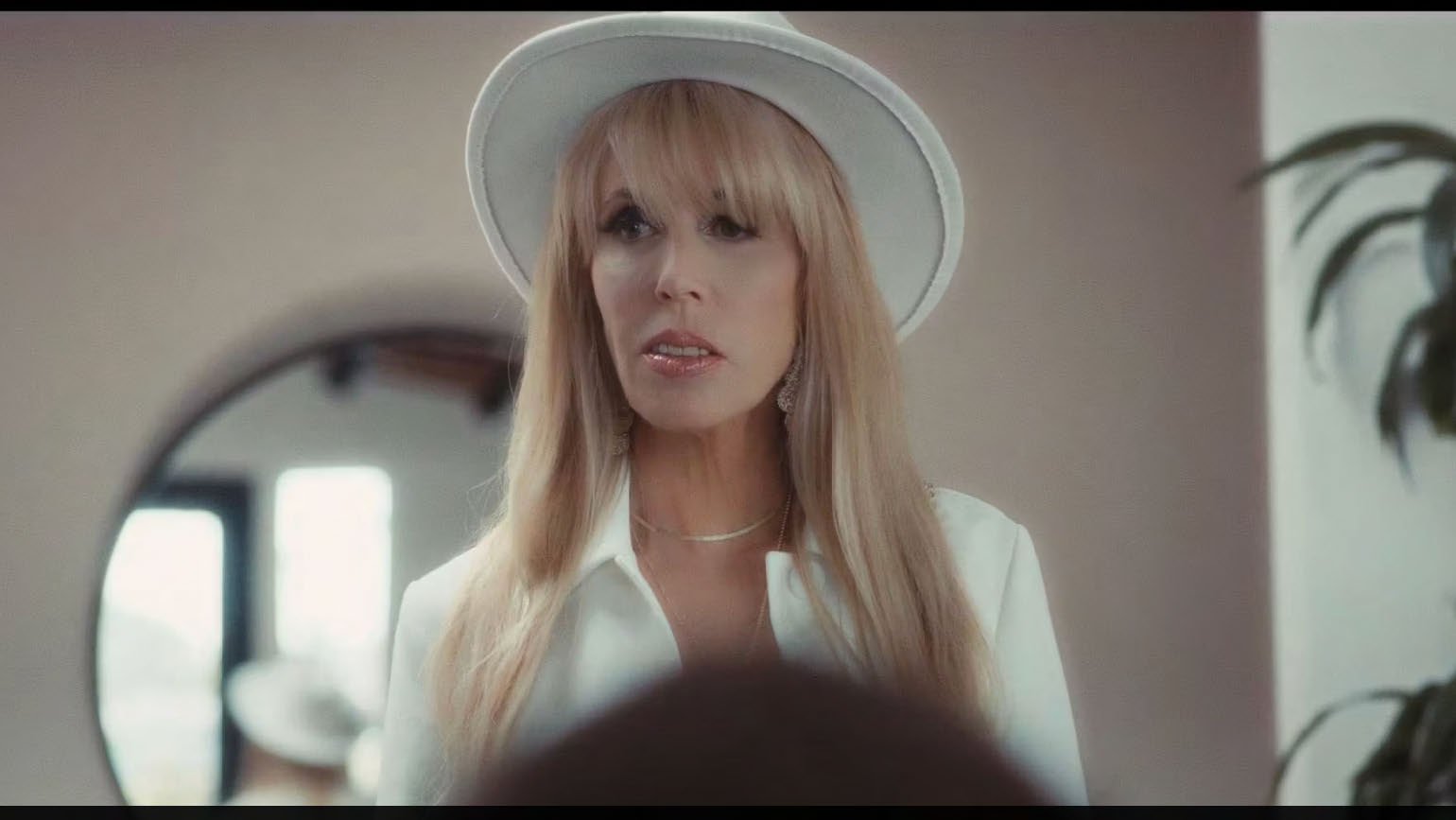 Woman with long blond hair wearing a white wide-brimmed hat and jacket, in front of a round mirror.