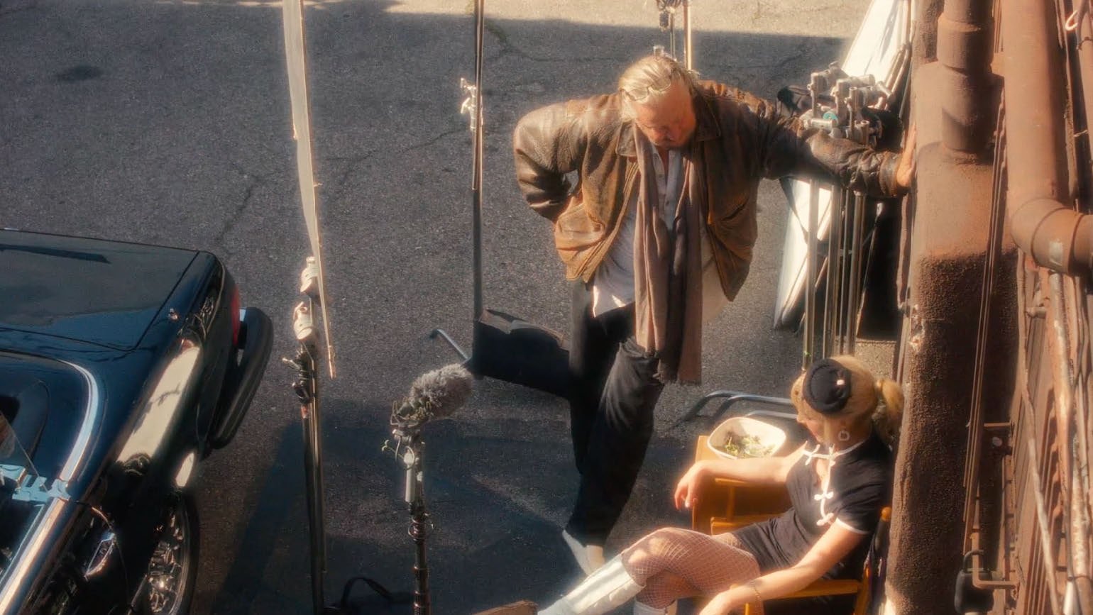 Filming scene with a person in a leather jacket and scarf leaning against a wall, a woman in a costume sitting in a director's chair, and a parked classic car nearby.
