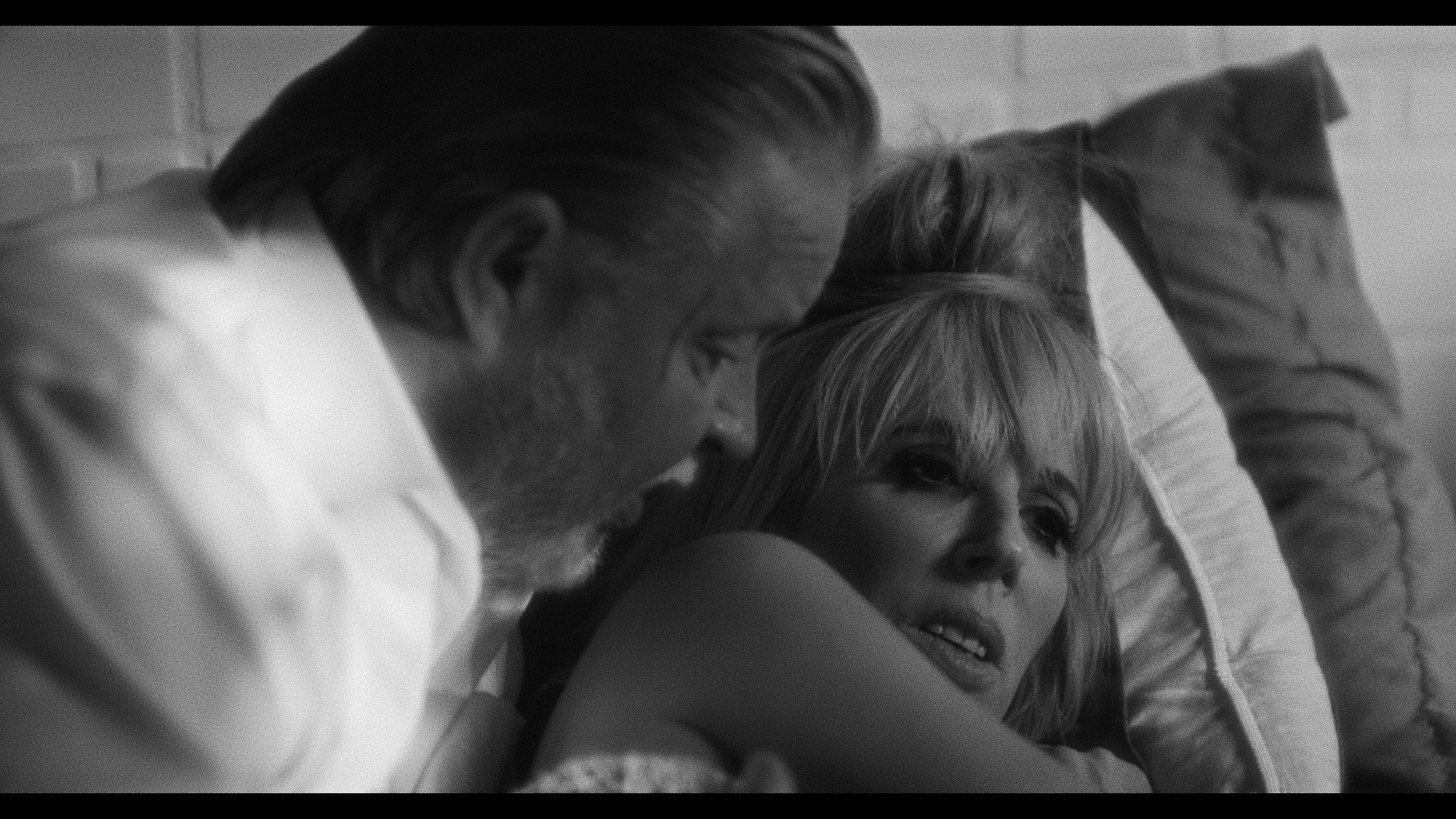 Black and white image of a man with facial hair leaning over a woman with blonde hair, both on a bed with pillows, appearing in a contemplative moment.