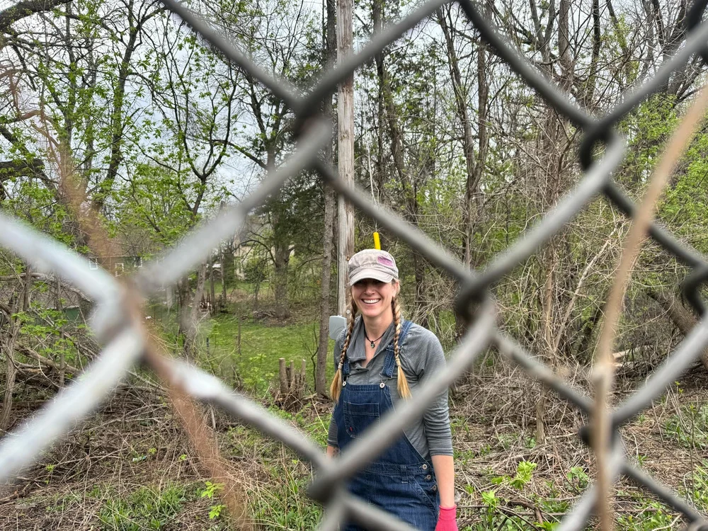 Looking though a chain-link fence at a woman clearing brush on the other side