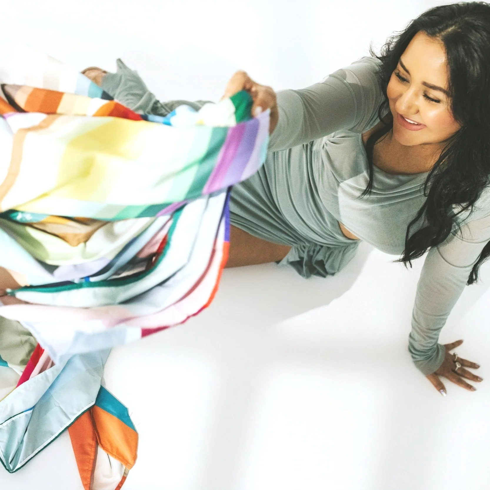 A woman with long dark hair, dressed in a gray top and shorts, kneeling on the floor, smiling while reaching into a colorful striped laundry basket.