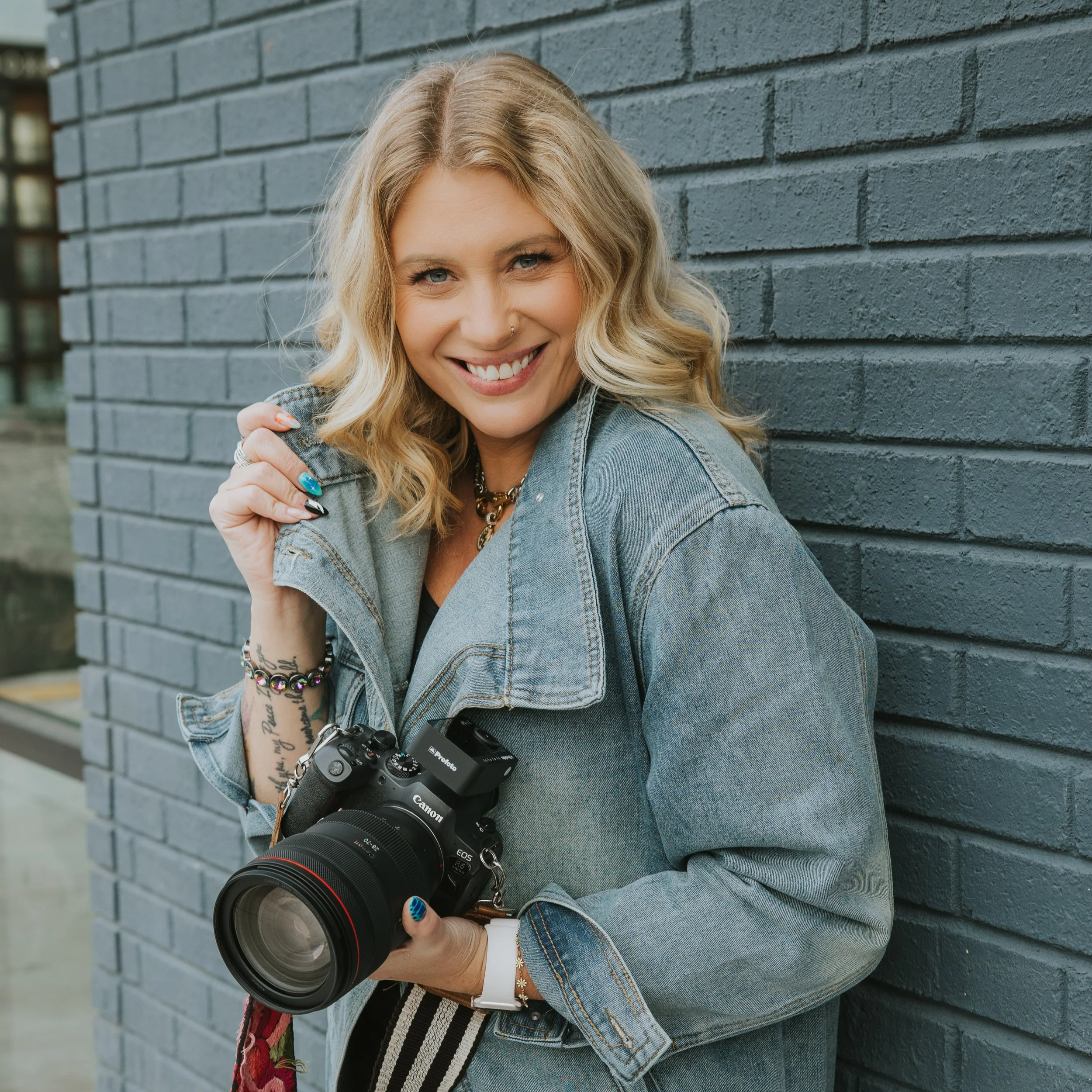 A young woman with blonde, wavy hair smiling while leaning against a dark grey brick wall. She holds a professional camera with a large lens and wears a denim jacket adorned with jewelry and nail art.