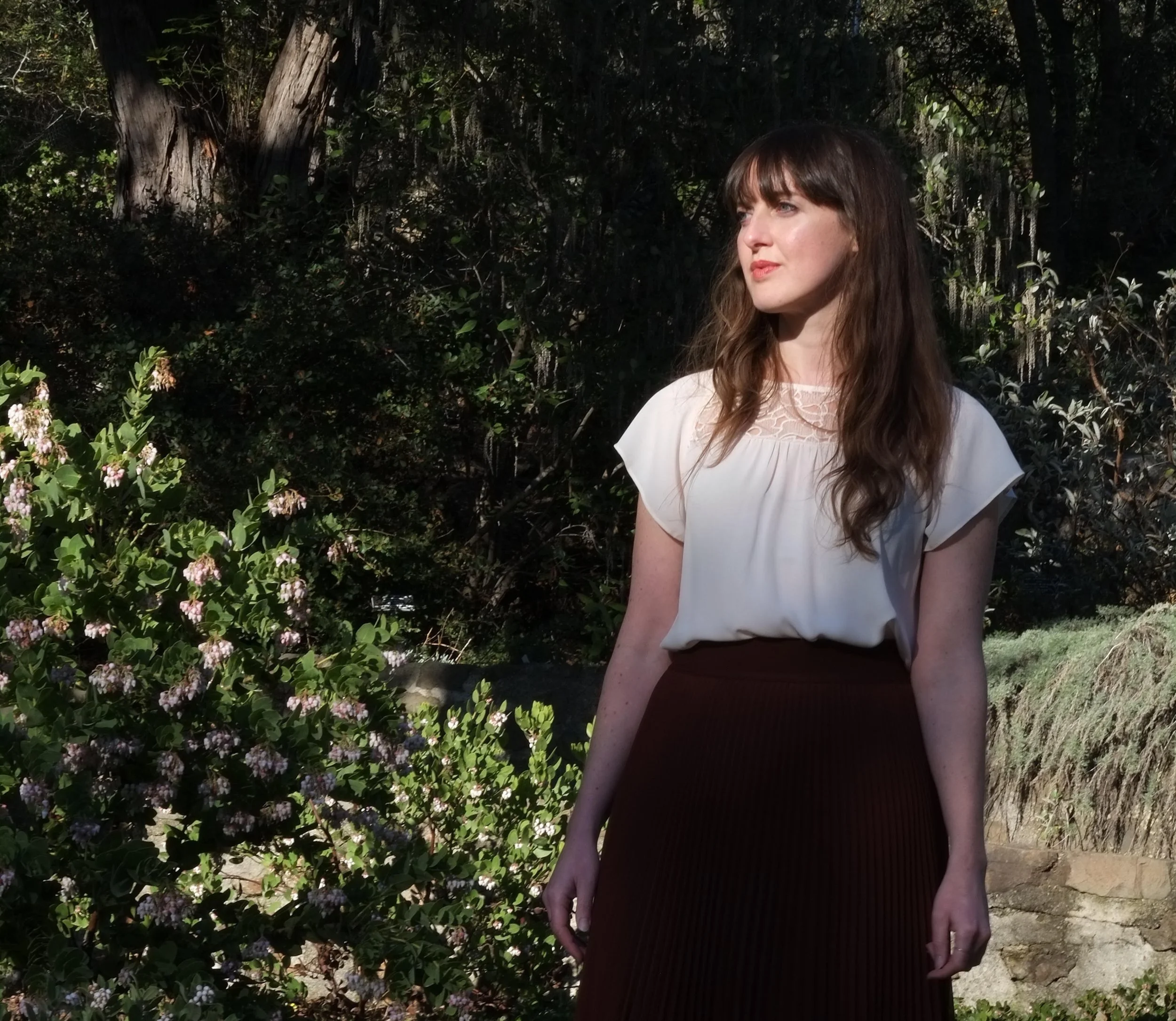 A woman with long brown hair wearing a white blouse and a maroon skirt stands outdoors near greenery and trees, looking to the right with sunlight illuminating her face.