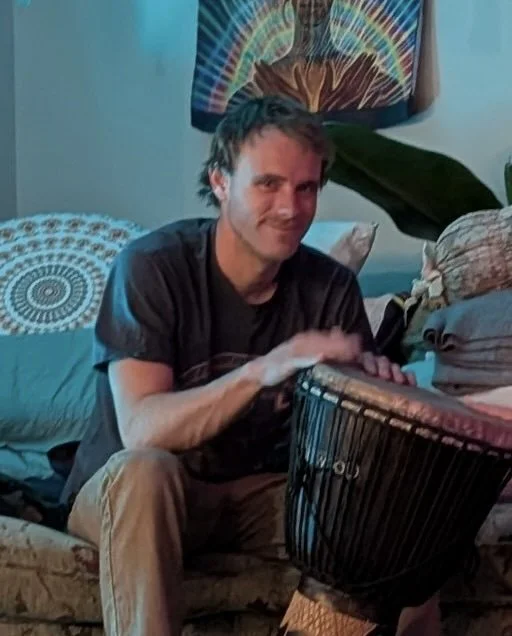 A man playing a djembe drum in a cozy living room, with vibrant wall art and decorative pillows.