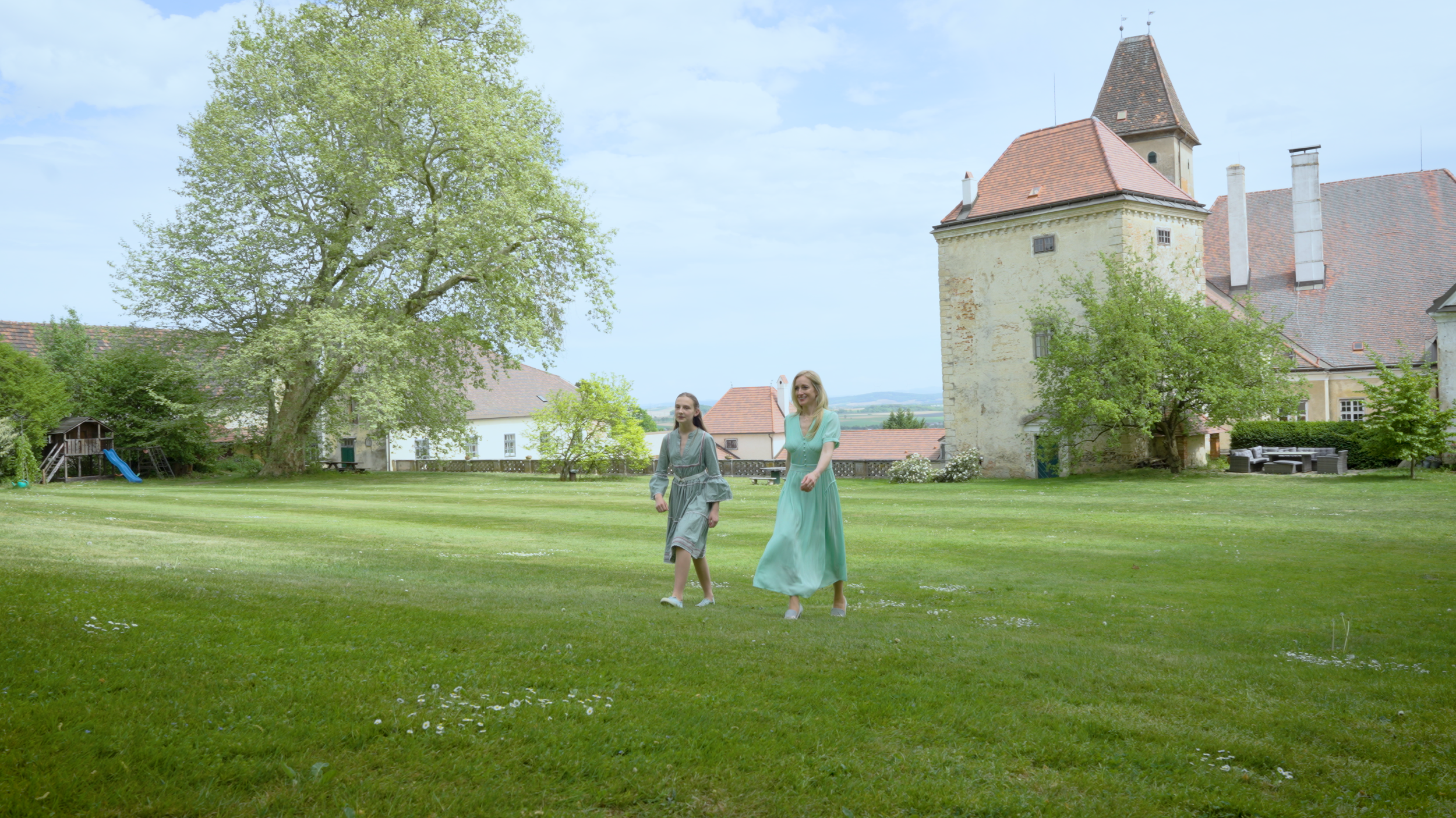 Zwei Frauen spazieren auf einem grünen Rasen vor einer alten Burg mit Türmen, umgeben von Bäumen und einem blauen Himmel.