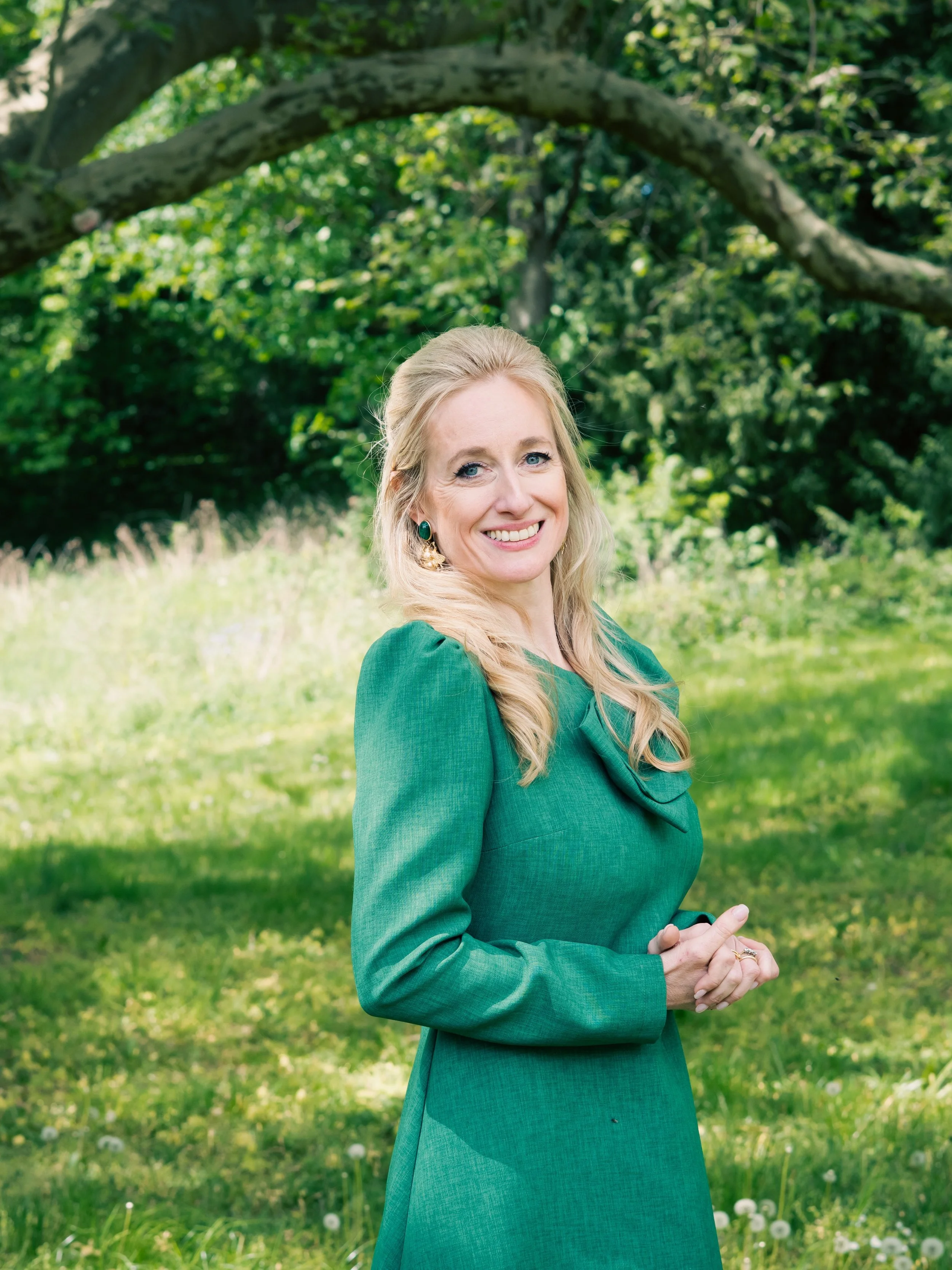 Eine blonde Frau in einem grünen Kleid steht vor einem Baum in einem Park mit grünem Gras und kleinen weißen Blumen.