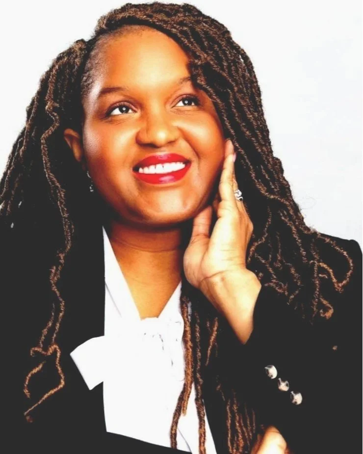 A Smiling African-American female Notary Professional with locks wearing a dark suit and white blouse, posing with hand on cheek against a white background.