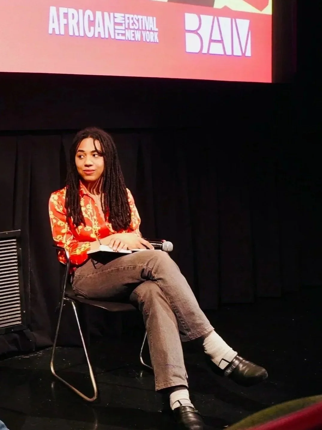 A woman with long dreadlocks sitting on a chair on stage at the African Film Festival New York, holding a microphone and notebook, wearing a red floral blouse, brown pants, and black shoes.