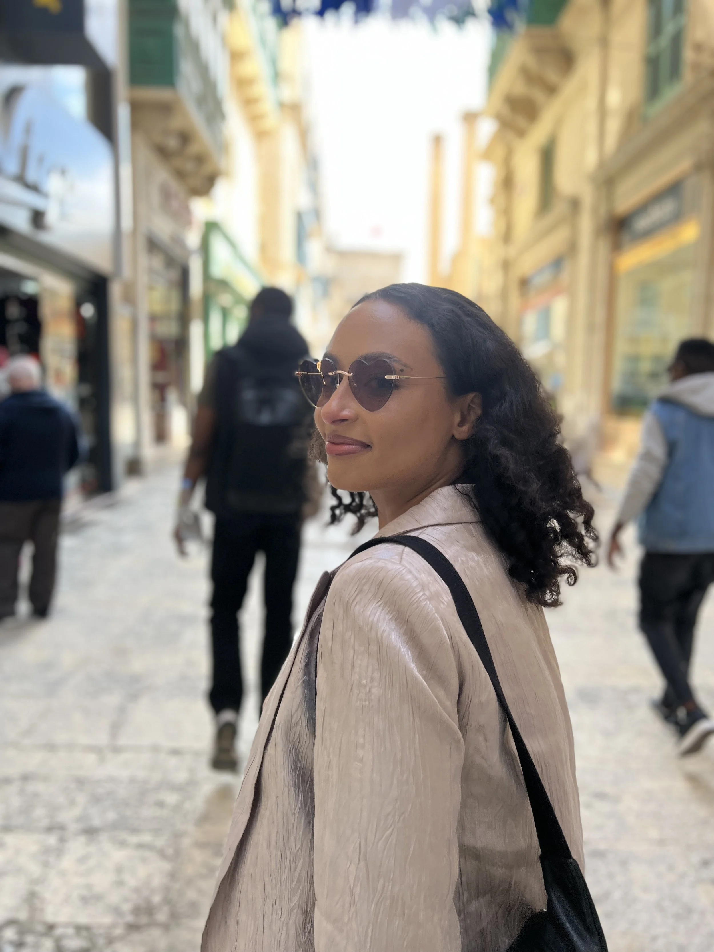 Image depicts Rachel Equity – a biracial woman with shoulder length curly brown hair –on a cobblestone street, looking back at the viewer over her left shoulder. She is wearing heart-shaped glasses and a silver blazer.