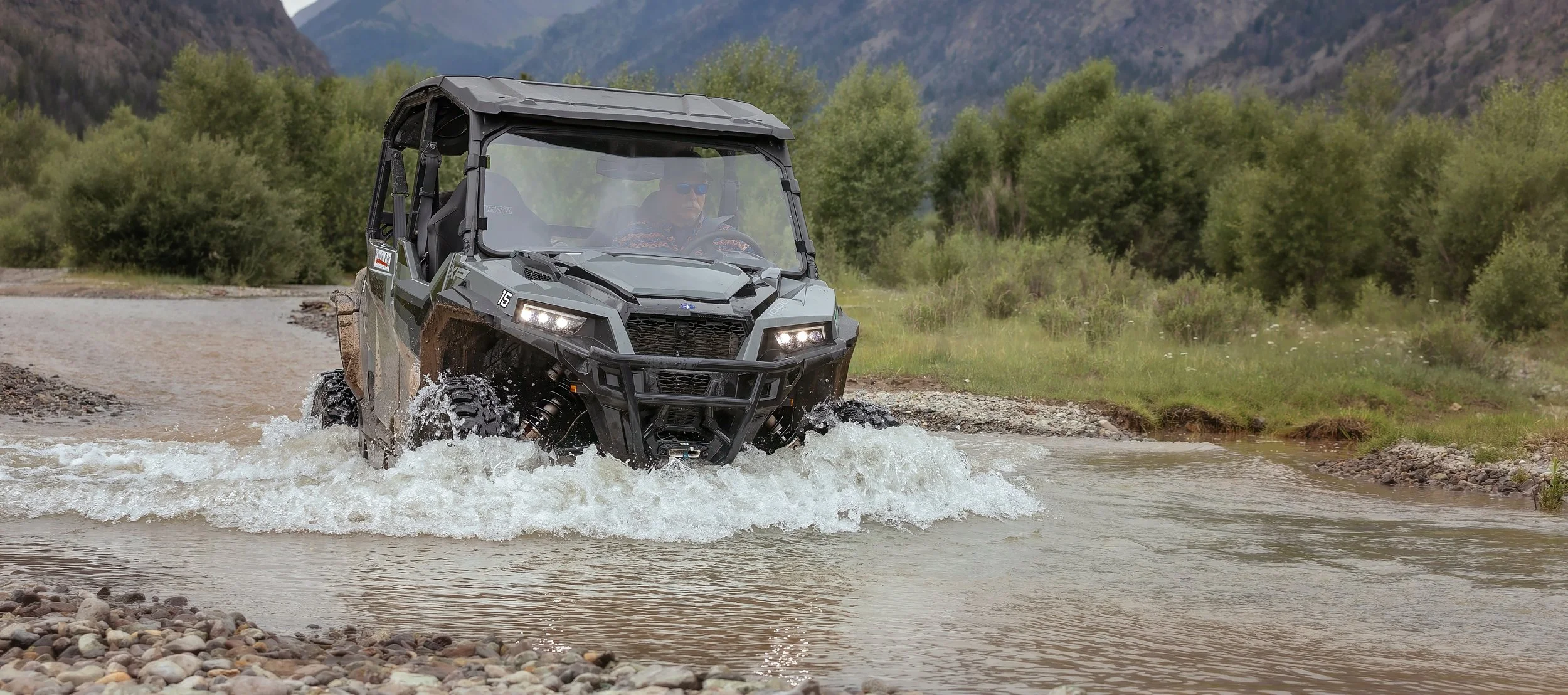 A person driving a black off-road utility vehicle through a river in a mountainous area with trees and hills in the background.