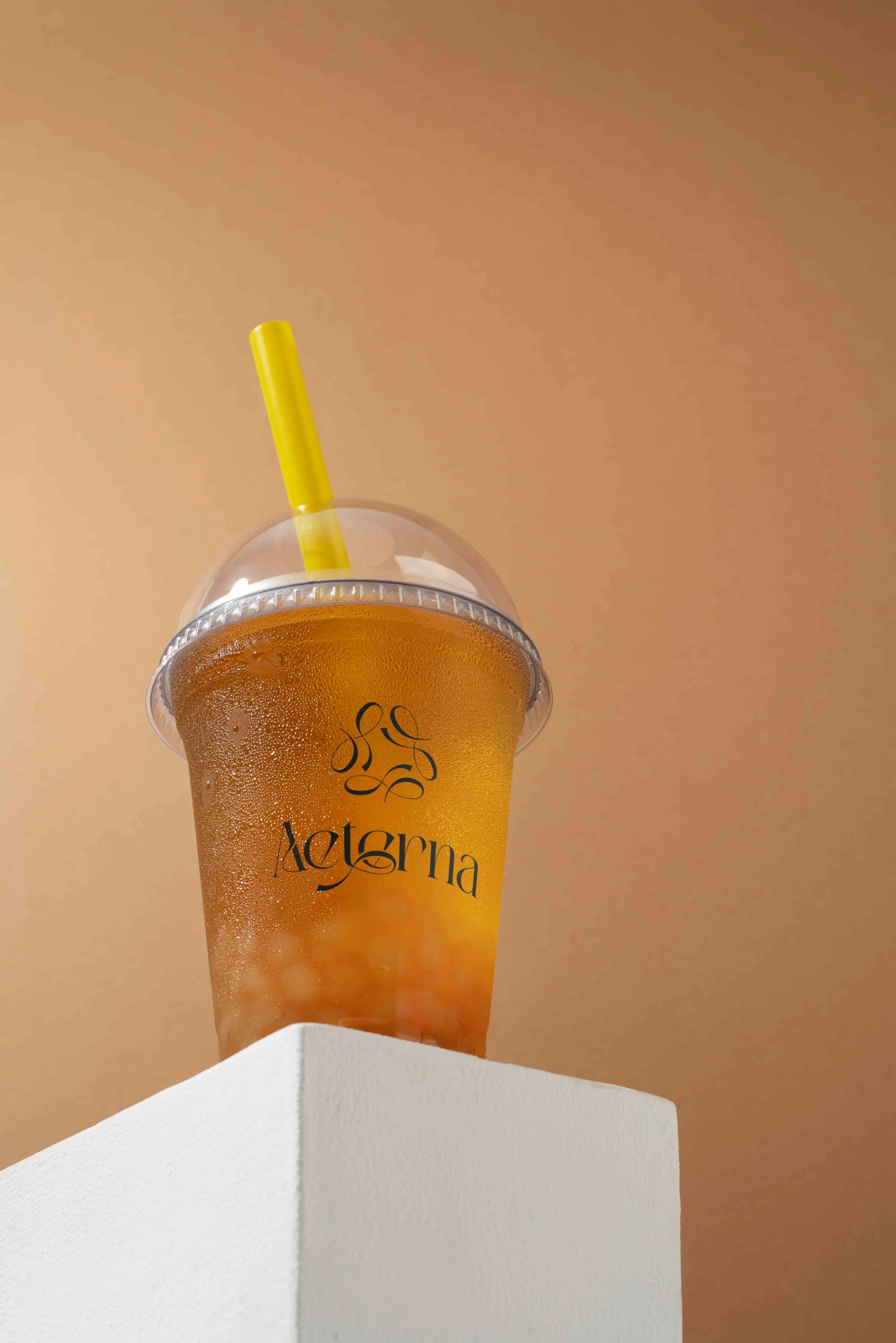 A plastic cup filled with a yellow-orange beverage with condensation on the outside, sitting on a white surface against a beige background. The cup has black decorative writing on it and a yellow straw.