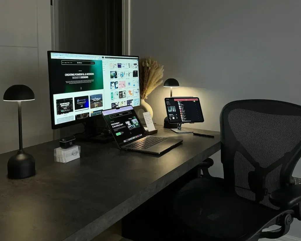 A workspace with a computer monitor, a laptop, a tablet, two small lamps, and a chair in front of a plain wall.