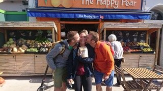 Three people smiling and hugging in front of a fruit stand at a market, with others in line behind them.