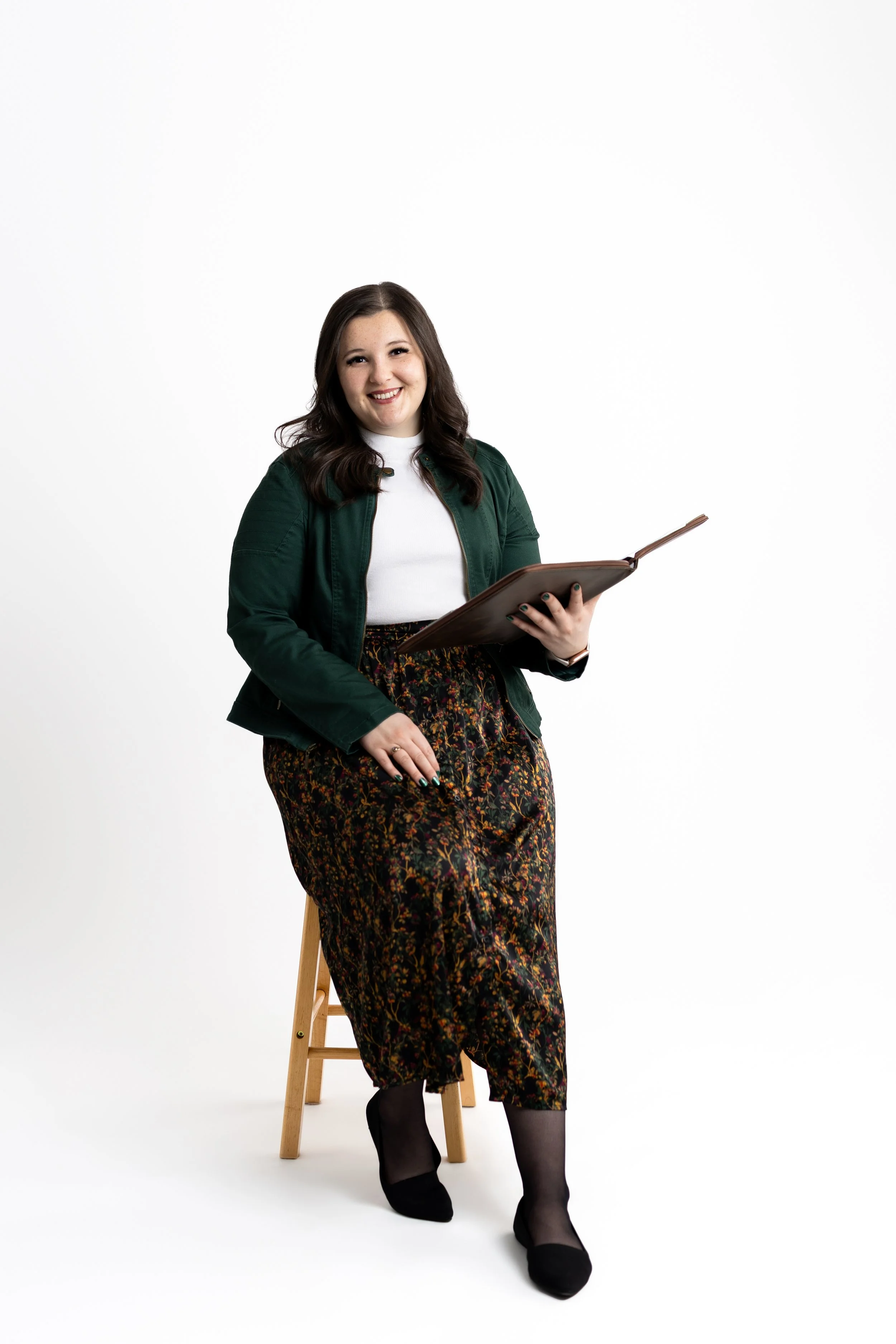 A woman with dark brown hair, wearing a dark green jacket, white top, and floral skirt, sitting on a wooden stool, holding a folder and smiling at the camera against a plain white background.