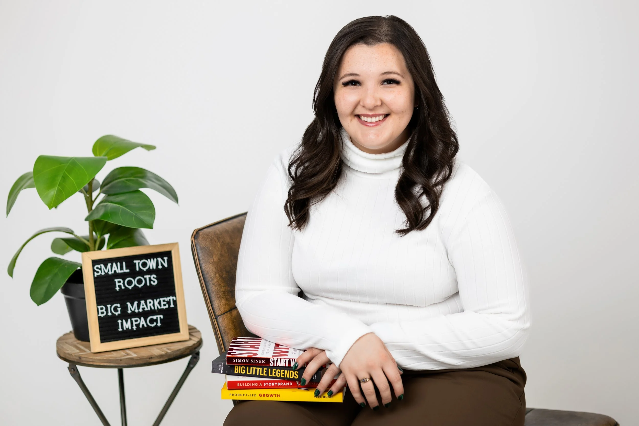 A woman with long dark hair and a white turtleneck sweater sitting on a chair, holding a stack of books, next to a small table with a potted plant and a sign that reads 'Small Town Roots, Big Market Impact.'