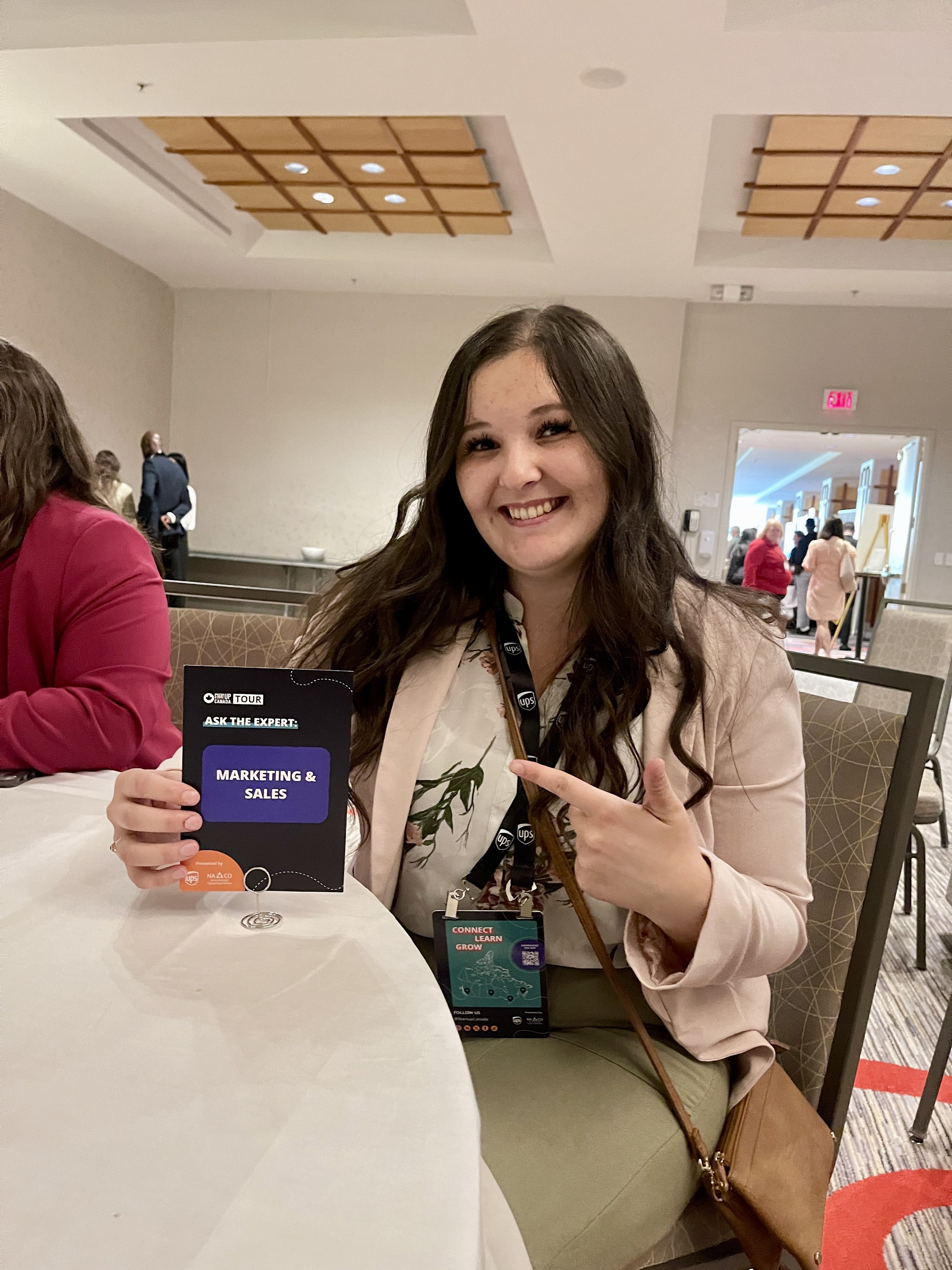 A woman with long dark hair, smiling and pointing to a card that says "Marketing & Sales," sitting at a table in a conference or event setting.