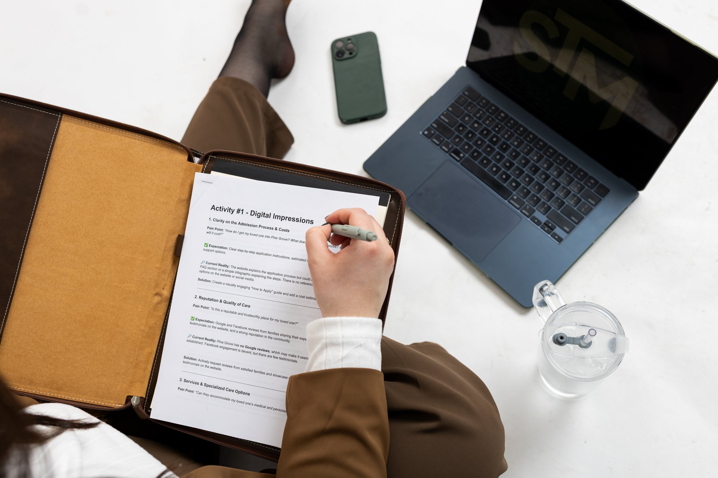 Person sitting at a white table working on a printed activity sheet titled 'Activity #1 - Digital Impressions' with a pen, a laptop, a smartphone, and a water bottle nearby.