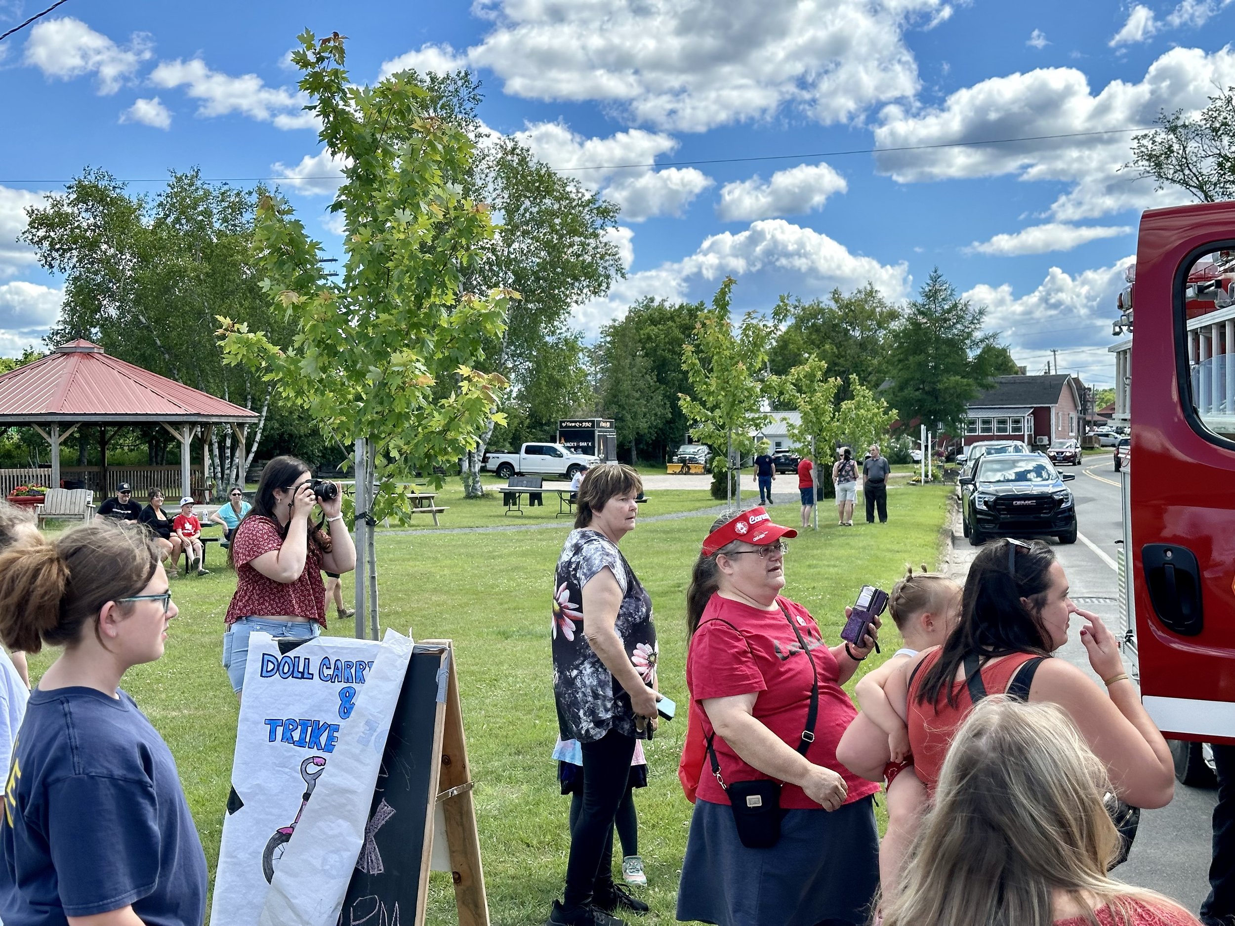 People gathered outdoors near a red fire truck, with some taking photos and others observing, on a sunny day with a blue sky and scattered clouds.