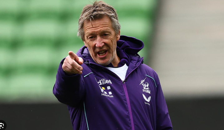 A man in a purple jacket pointing and speaking, standing outdoors.