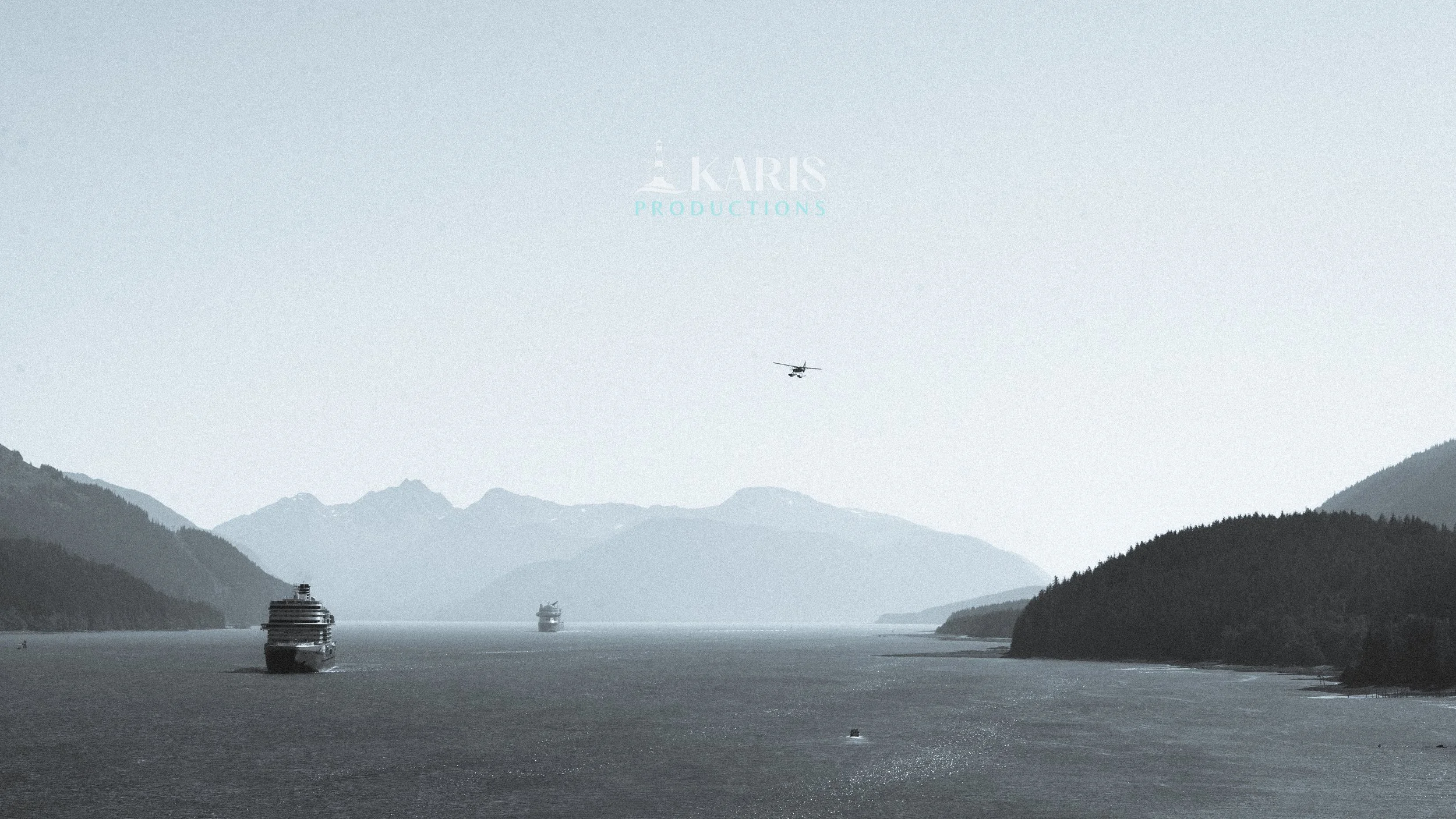 A scenic view of a waterway with two large boats, surrounded by forested mountains in the background, and a helicopter flying in the sky. Juneau, Alaska. Karis Productions copyright 2025