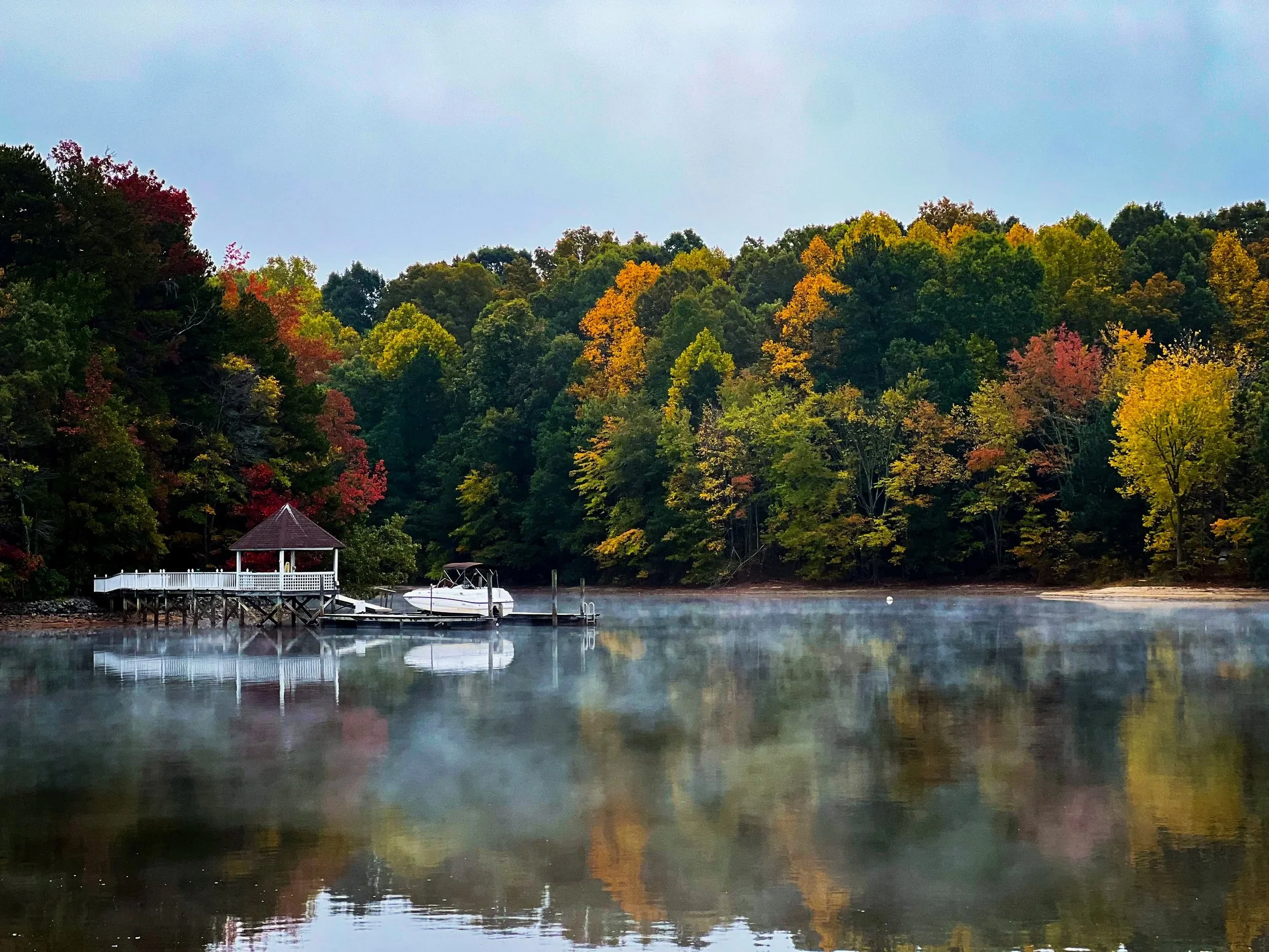 A lake with a white dock and boat reflecting on the water, surrounded by trees with autumn foliage, and a gazebo or small house at the shoreline.