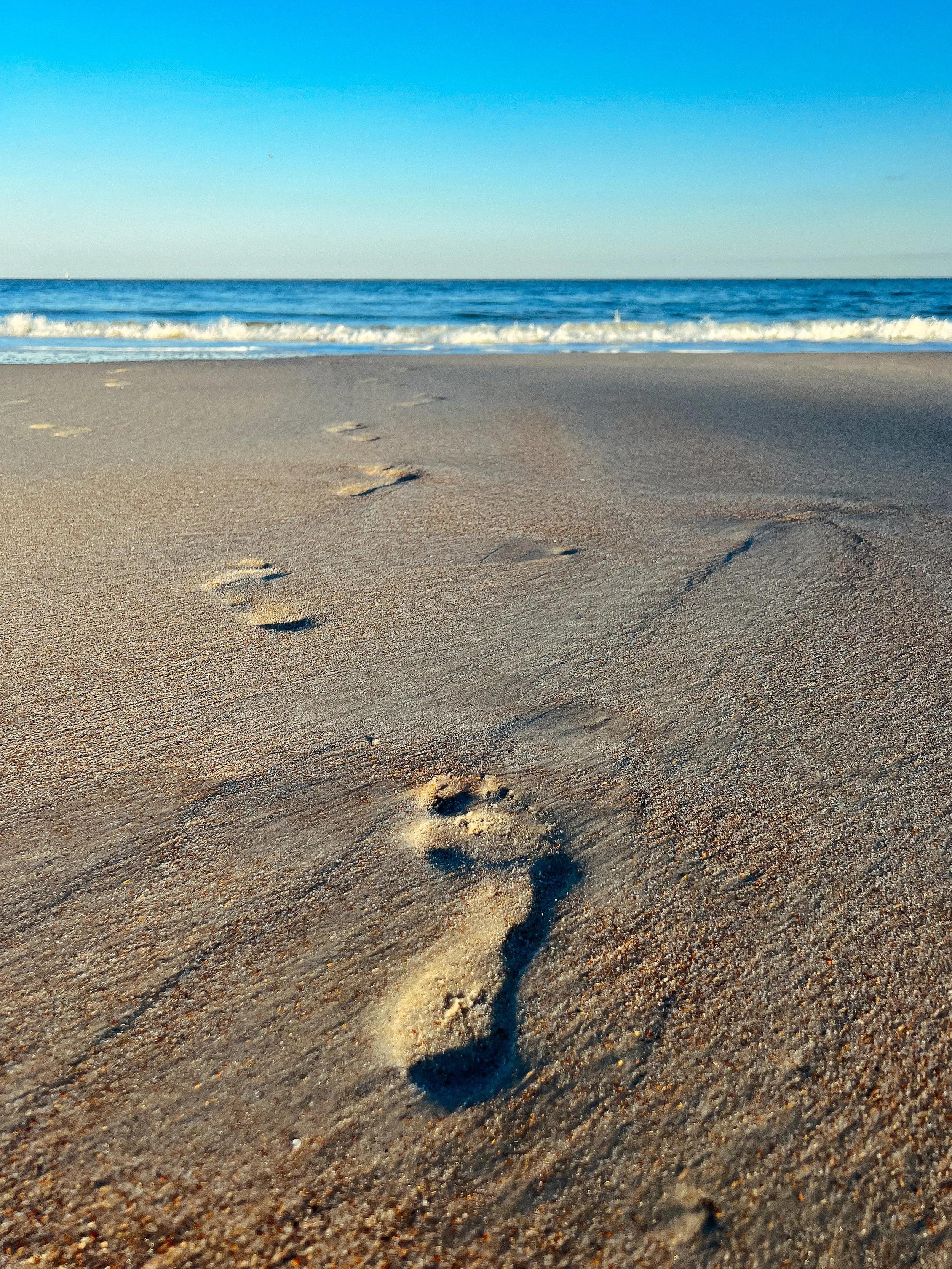 Footprints leading towards the ocean on sandy beach with waves and blue sky.
