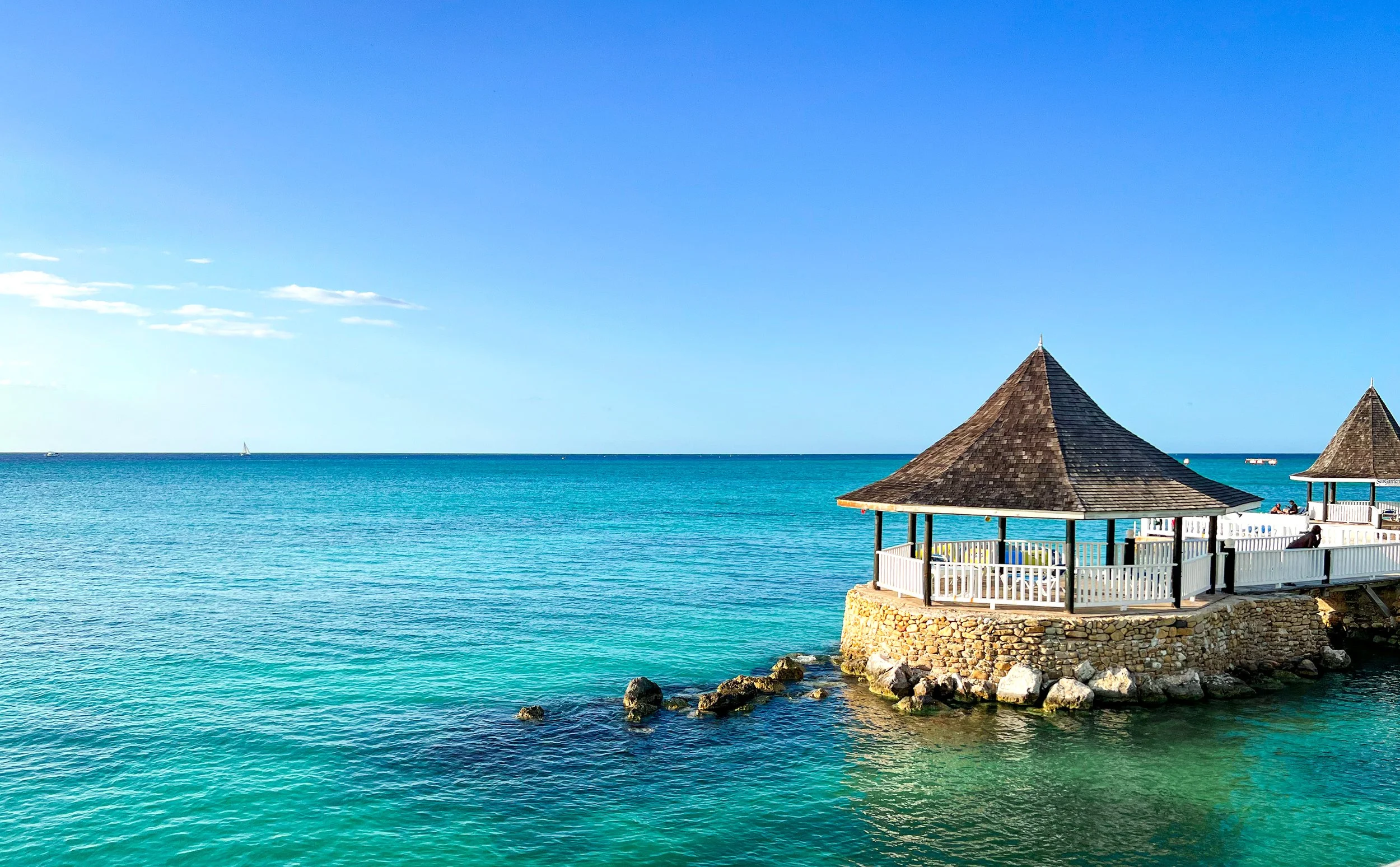 A clear blue ocean with a sailboat in the distance and a wooden gazebo on rocks at the shore under a bright sky.