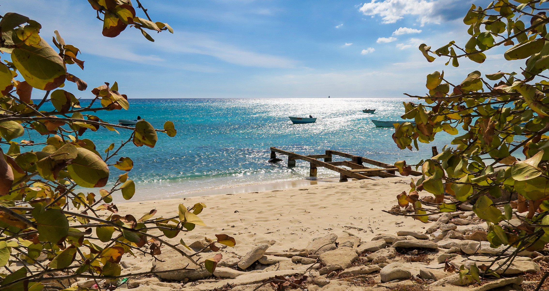 View of a sandy beach with a wooden pier, turquoise water with boats, and a blue sky with a few clouds, framed by leaves and branches in the foreground.