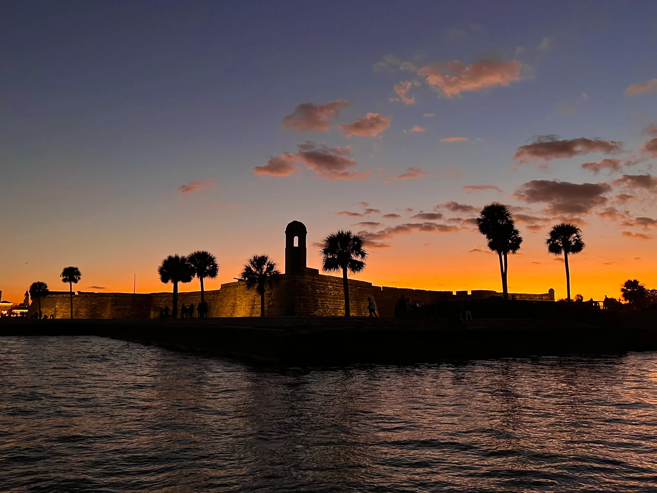 A sunset over a body of water with a silhouette of historic fort and palm trees, pink and orange clouds in the sky.