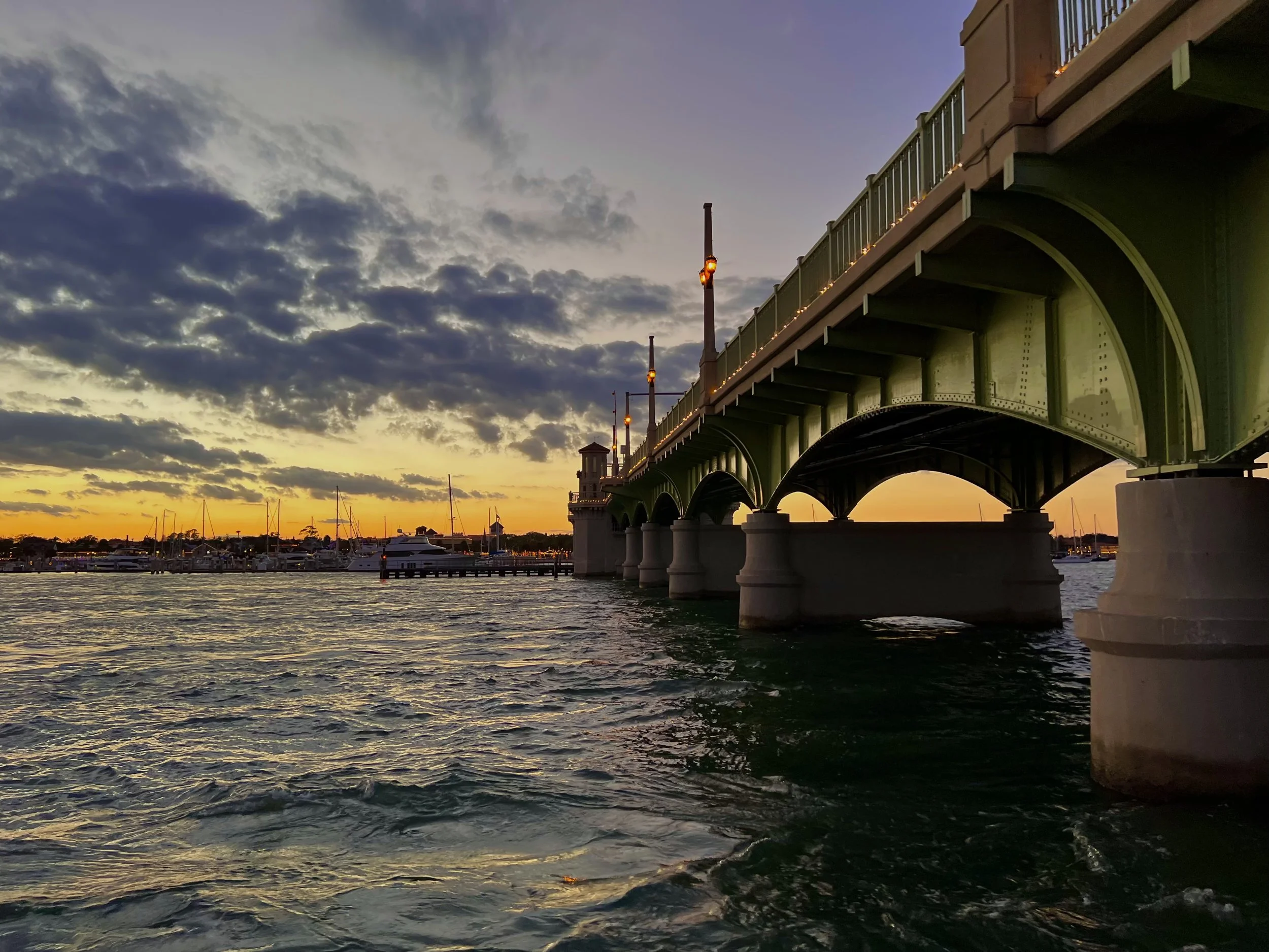 A view of a bridge over a body of water during sunset, with boats docked in the background and the sky partly cloudy.