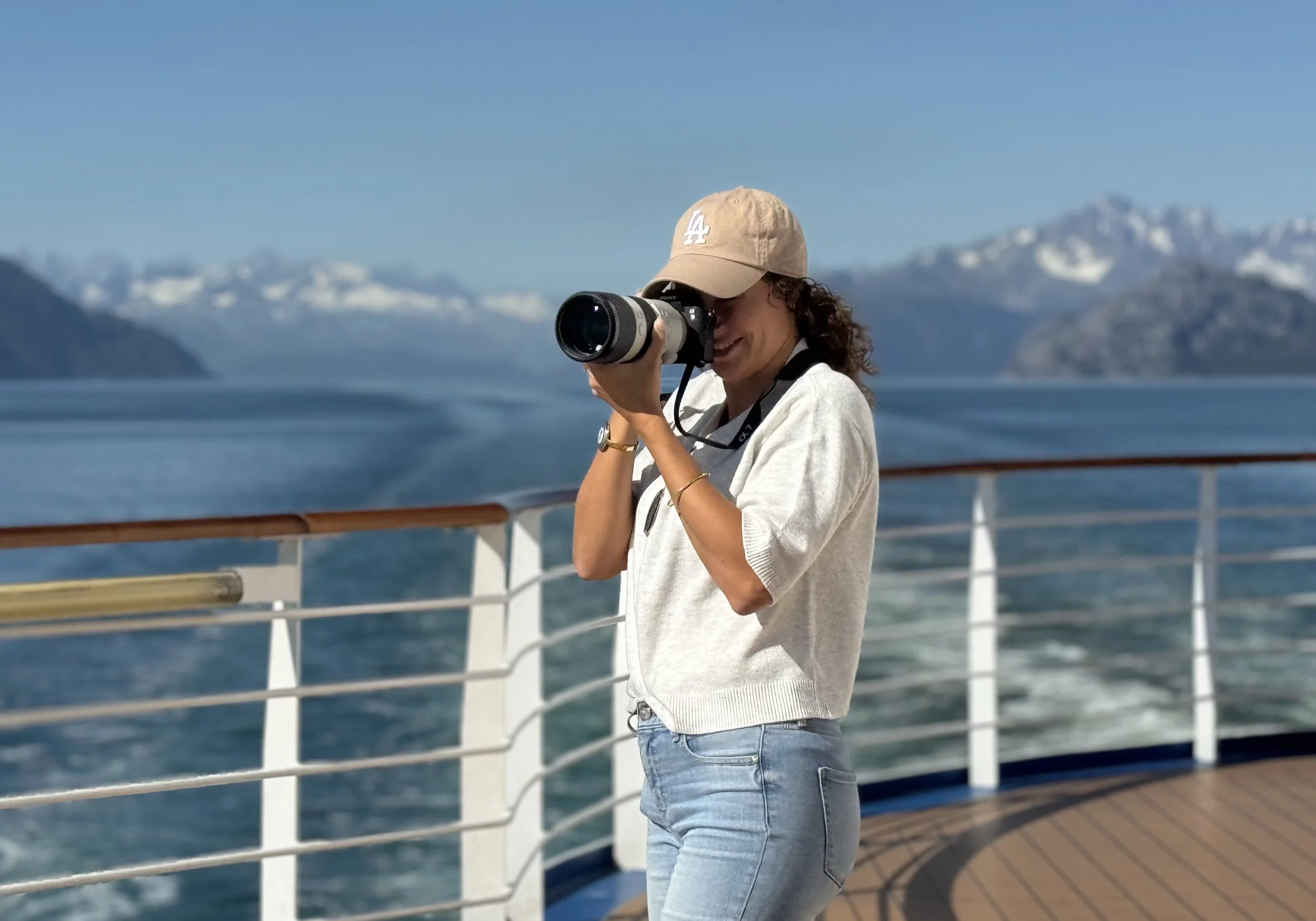 A woman with curly hair takes a photograph with a camera on a boat, with snow-capped mountains and a clear blue sky in the background.