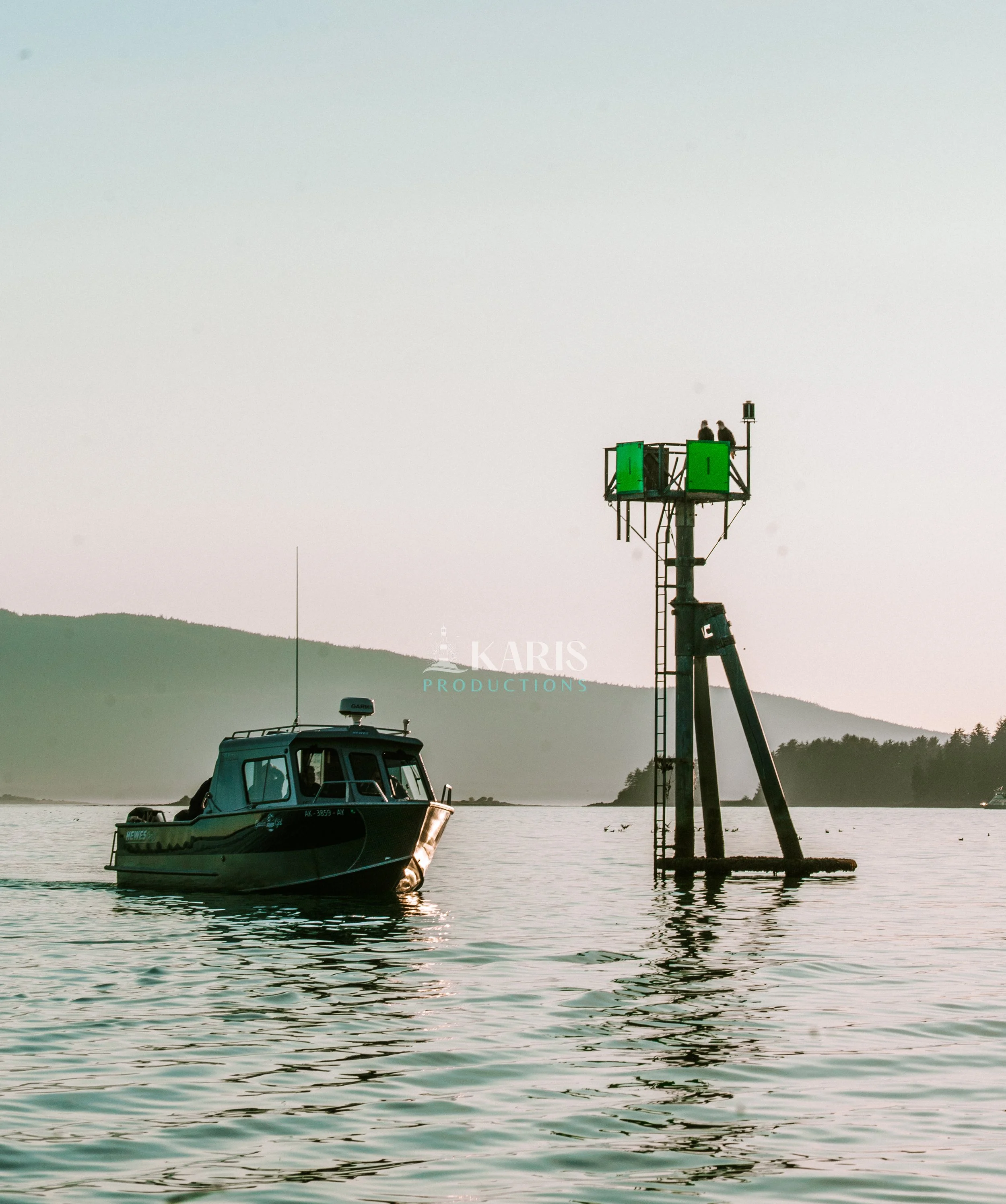 A boat on the water near a structure with two bald eagles sitting at the top, and hills in the background under a clear sky. Juneau, Alaska. Karis Productions copyright 2025