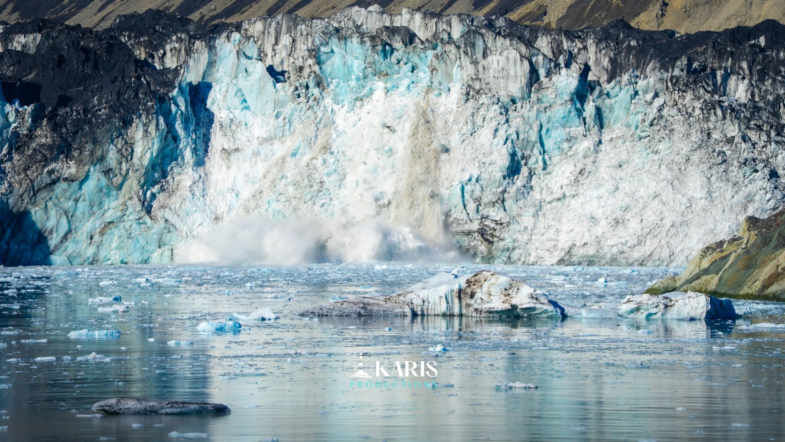 A glacier calving into the ocean, with ice falling from the glacier's edge and breaking into smaller ice chunks in the water. Karis Productions copyright 2025