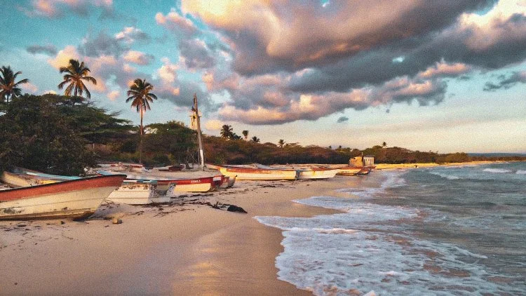 Boats resting on a sandy beach with palm trees and a partly cloudy sky in the background during sunset or sunrise.