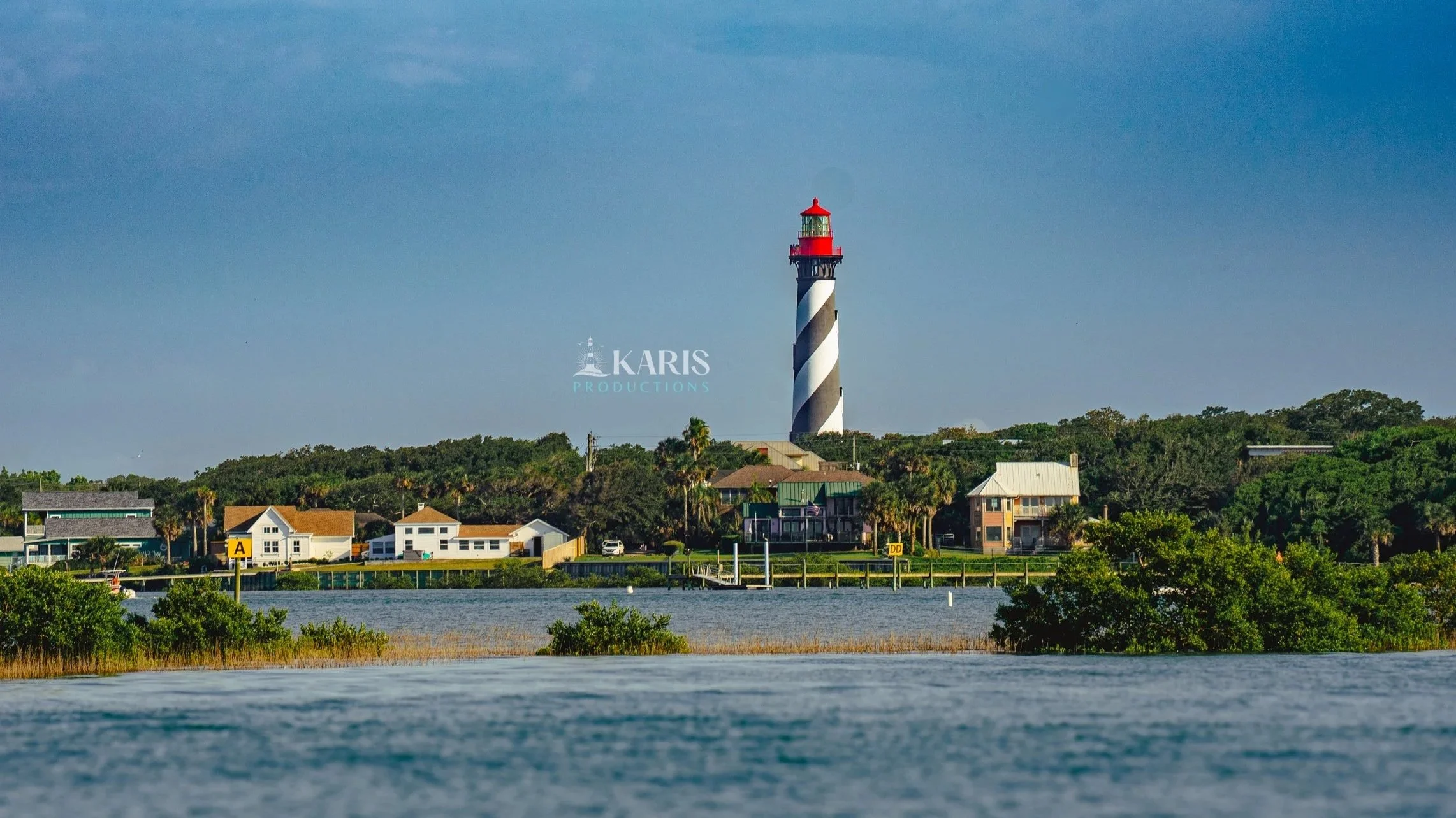 A coastal scene with a waterway in the foreground, residential houses and greenery in the middle ground, and a lighthouse with a red top and black and white spiral stripes in the background.