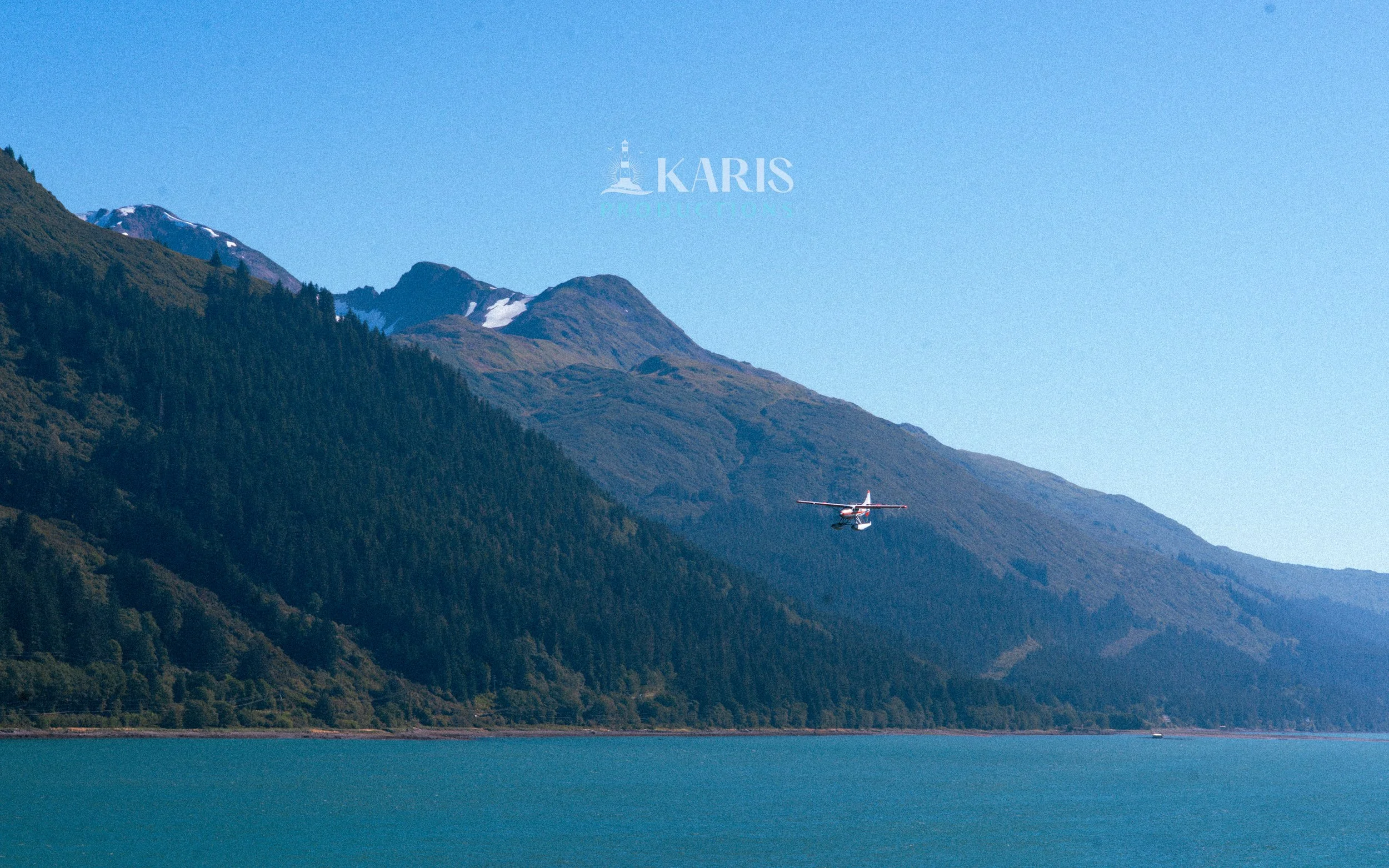 A small airplane flying over a body of water with green mountains in the background, clear blue sky in Juneau, Alaska. Karis Productions copyright 2025