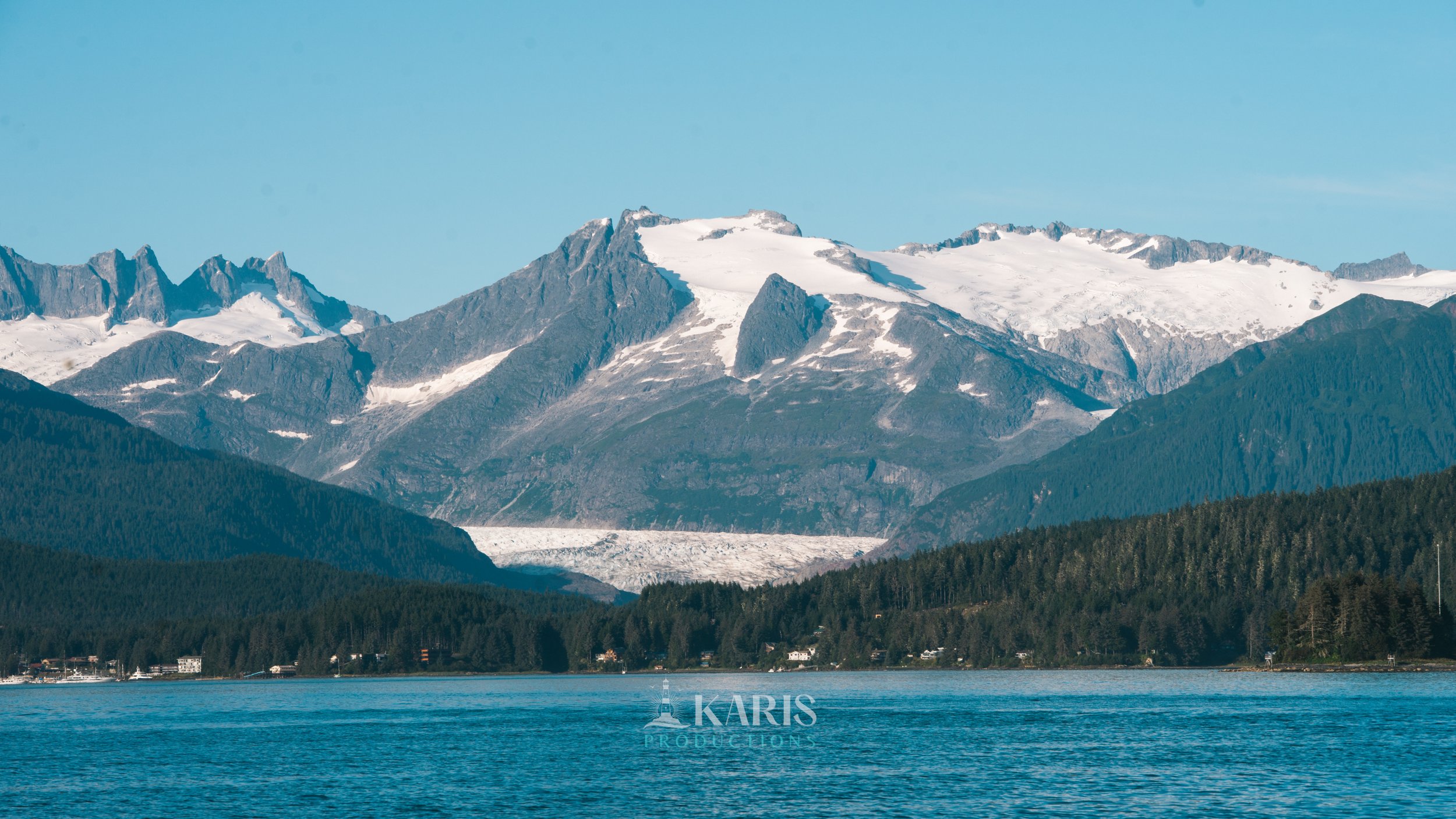 Snow-capped mountains towering over a forested landscape with a body of water in the foreground under a clear blue sky. Karis Productions copyright 2025