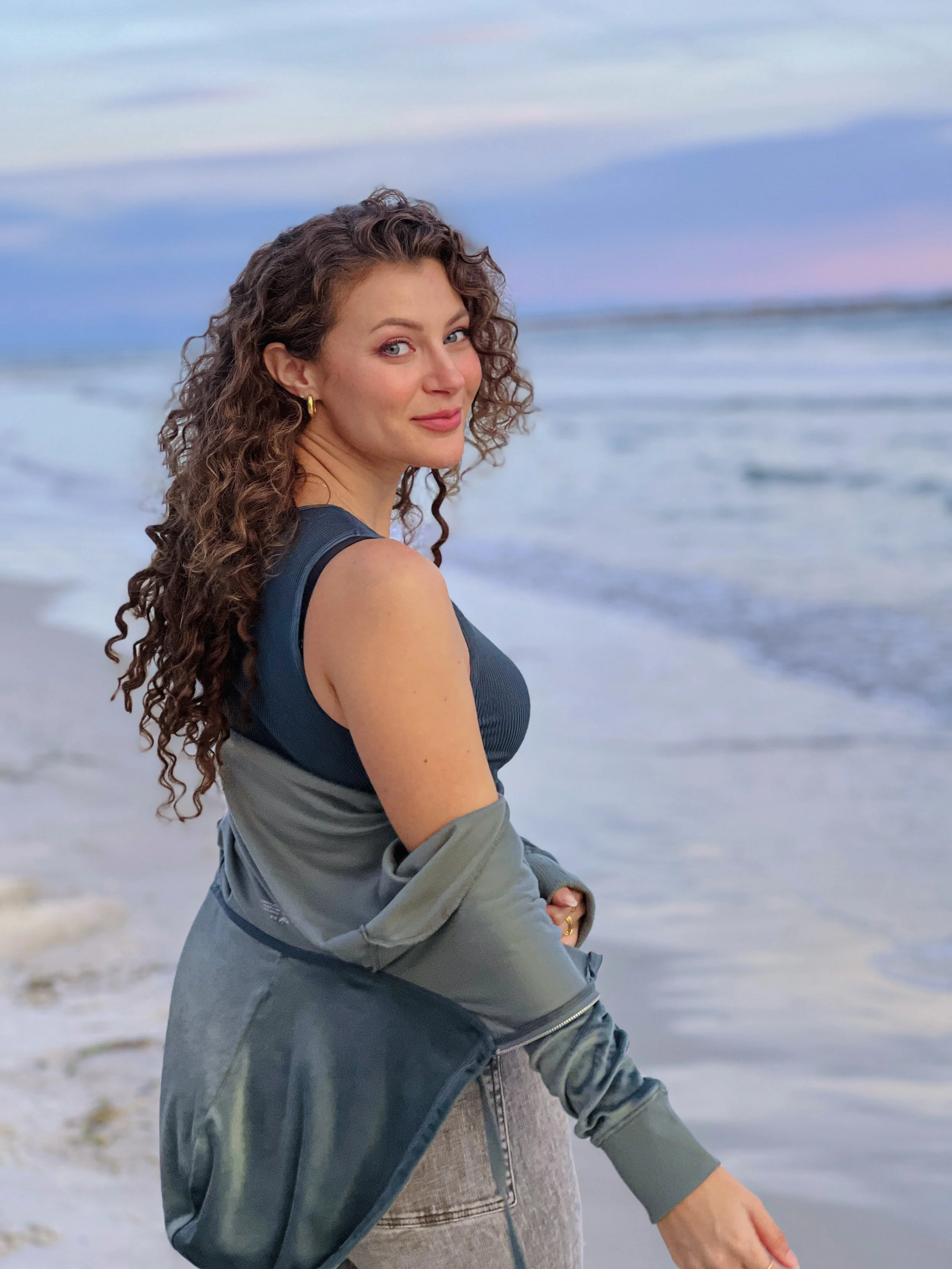 A woman with curly brown hair and blue eyes, wearing a sleeveless dark top, a gray jacket draped over her arm, and gold earrings, standing on a beach with ocean waves and a pastel-colored sky in the background.