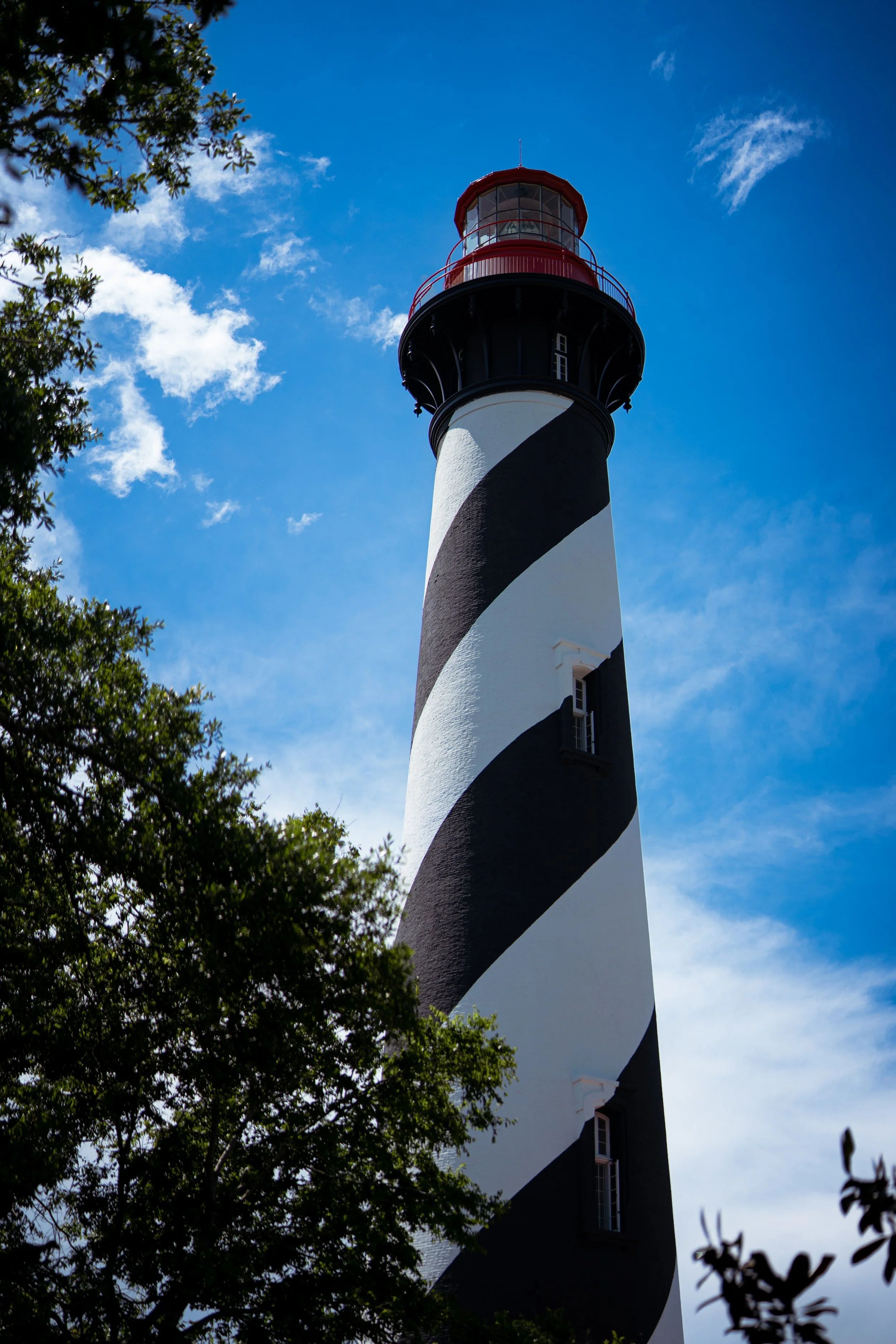 A lighthouse with black and white diagonal stripes on its tall tower, with a red balcony and lantern room at the top, against a blue sky with some clouds, partly obscured by trees in the foreground.