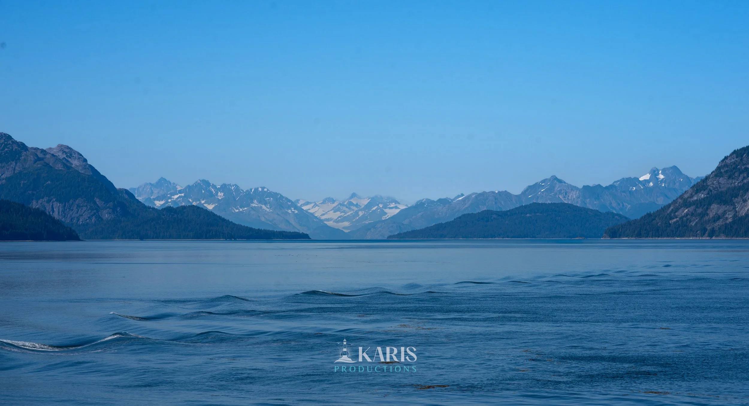 Scenic view of a calm body of water with mountains and snow-capped peaks in the background under a clear blue sky. Juneau, Alaska. Karis Productions copyright 2025